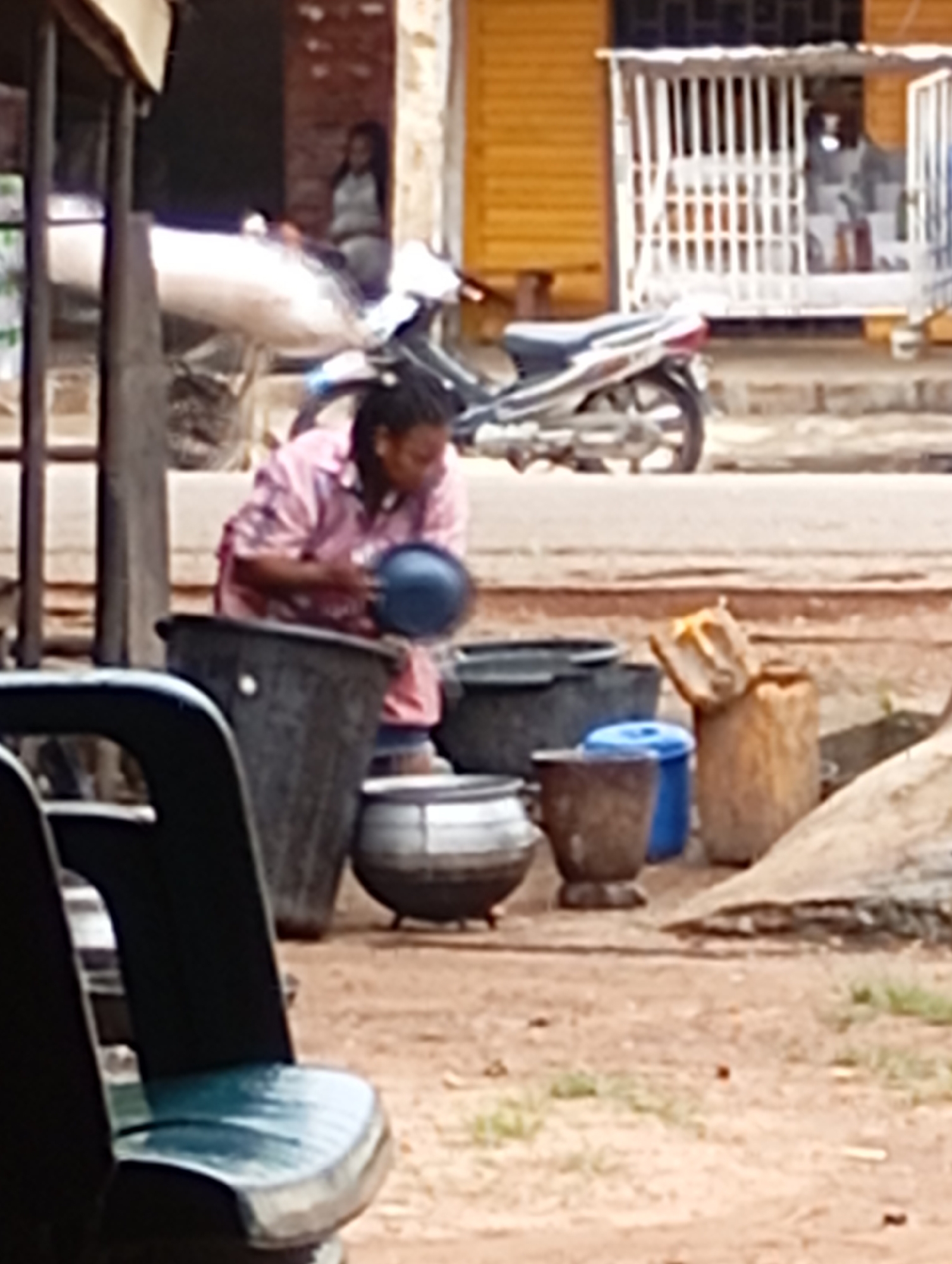 The image depicts an outdoor scene in Shendam, Nigeria, with a focus on a person and various objects. A woman, with dark braided hair, is wearing a pink patterned shirt and is holding a dark bowl over a large black tub. In the background, a motorcycle is parked. To the right of the woman, there are several large pots and containers, including a blue container and a large, light brown cylindrical object that appears to be made of wood or a similar material. Another large, dark pot is visible to the left of the woman. The ground is a mixture of dirt and scattered vegetation. A portion of a dark green chair or bench is in the foreground, partially obscuring the lower left of the scene. The overall environment suggests a marketplace or an area where goods are being prepared or stored.