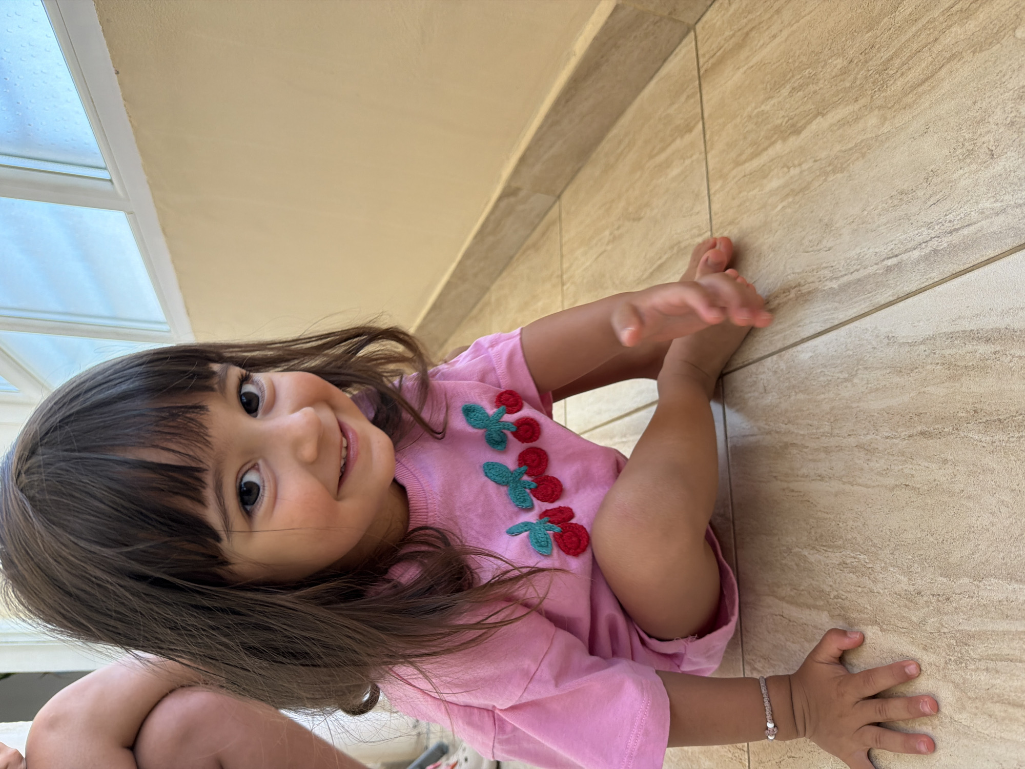 A young child with dark hair and bangs is sitting on a tiled floor, looking at the camera with a smile. The child is wearing a pink t-shirt with embroidered cherries. To the left of the child, another person's legs and knee are visible, suggesting they are nearby. The background shows a wall and a window with blue sky visible through it. The overall scene appears to be indoors, possibly in a home or a building with an outdoor area. The location of Burgas, Bulgaria is not determinable from the image.