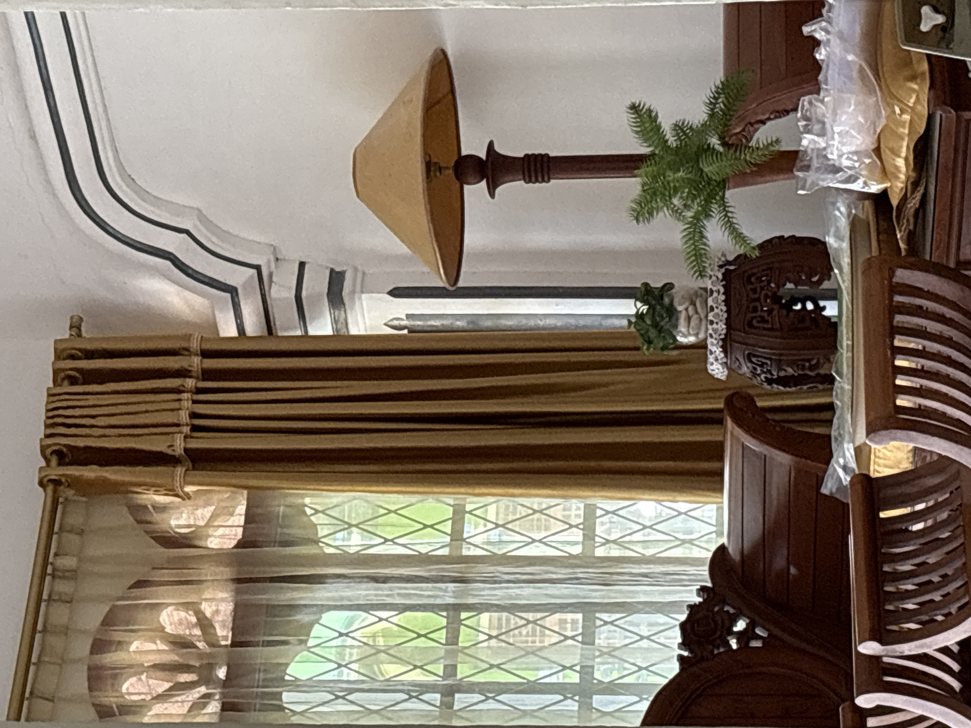 The image shows an interior scene in what appears to be a living room. The room features ornate wooden furniture, including a sofa with carved detailing and slatted seating. A tall floor lamp with a conical, tan lampshade stands to the right, positioned next to a small potted plant on a decorative wooden stand. A sprig of greenery, possibly a decorative branch, is placed in the base of the lamp. Light streams through a diamond-paned window covered by sheer and heavier drapes in shades of tan and gold. The architectural elements of the room include a decorative archway with dark trim. Plastic-wrapped items are visible on the furniture, suggesting the space may be in transition or storage. The scene is brightly lit, with natural light filtering through the window. The photograph is taken from a medium distance, capturing a good portion of the room's decor. The location is Uleepulo, Indonesia.