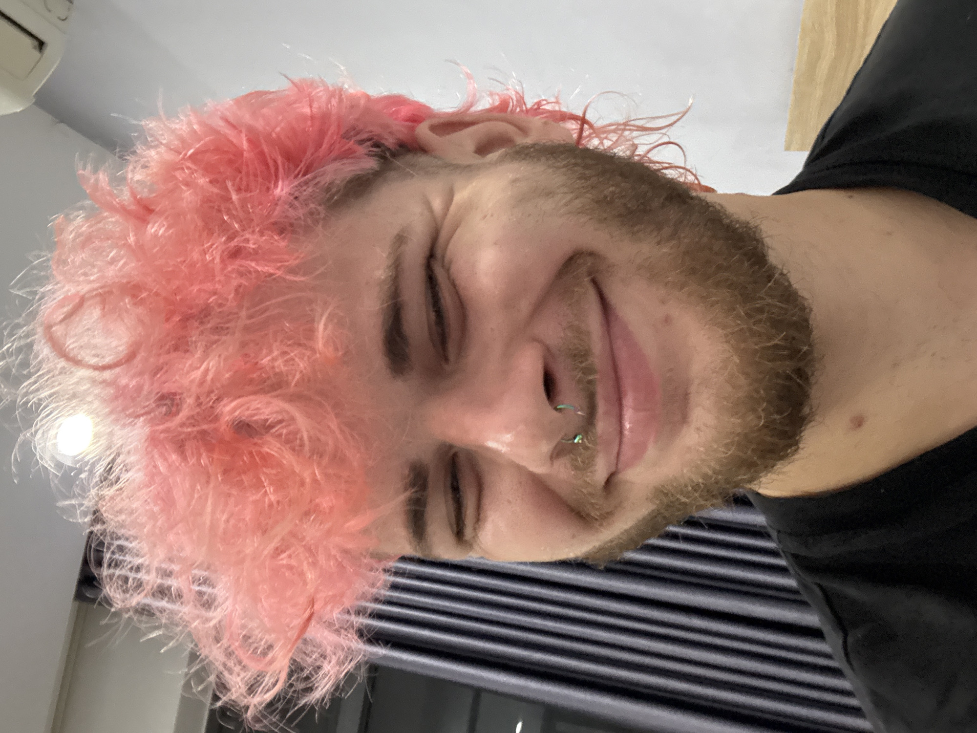 A person with pink hair and a beard is smiling. They have a nose ring. The person is wearing a black shirt. The background features what appears to be a window or blinds. There is no indication the photo was taken in Đà Nẵng, Vietnam.