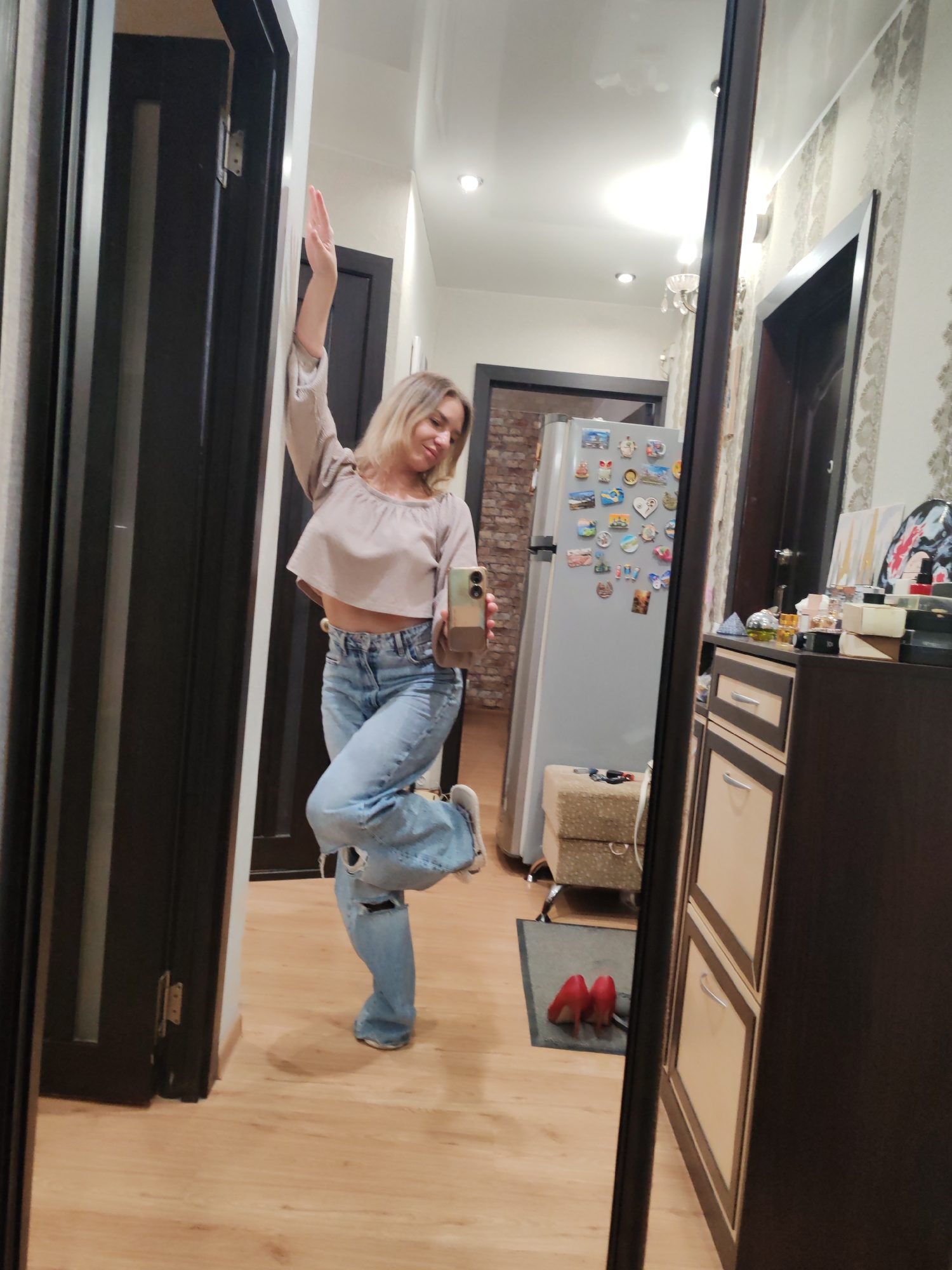 A young woman with blonde hair poses in front of a full-length mirror indoors. She wears a cropped beige long-sleeved top and ripped blue jeans, with one leg bent. She holds a smartphone in her right hand, presumably taking a selfie. In the reflection behind her, a hallway is visible, featuring a refrigerator covered in magnets, a storage ottoman, and a glimpse of a brick-patterned wall. Two red high-heeled shoes rest on the floor near the base of the mirror. The setting appears to be a modern apartment. There is no explicit evidence indicating that this photo was taken in the city of Petropavlovsk-Kamchatsky, Russia.