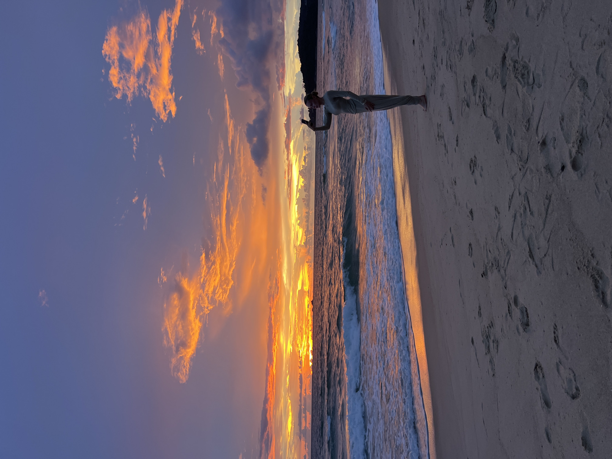 A lone woman stands on a sandy beach, facing a vibrant sunset over the ocean. The sky is a dramatic display of orange, pink, and purple hues, with scattered clouds catching the last rays of light. The ocean is choppy with white-capped waves rolling towards the shore, reflecting the colorful sky. The woman is wearing a long-sleeved jumpsuit and is holding up a mobile phone, seemingly taking a selfie. Footprints are visible in the sand leading up to her. The overall scene captures a moment of tranquil beauty and solitude at the end of the day.