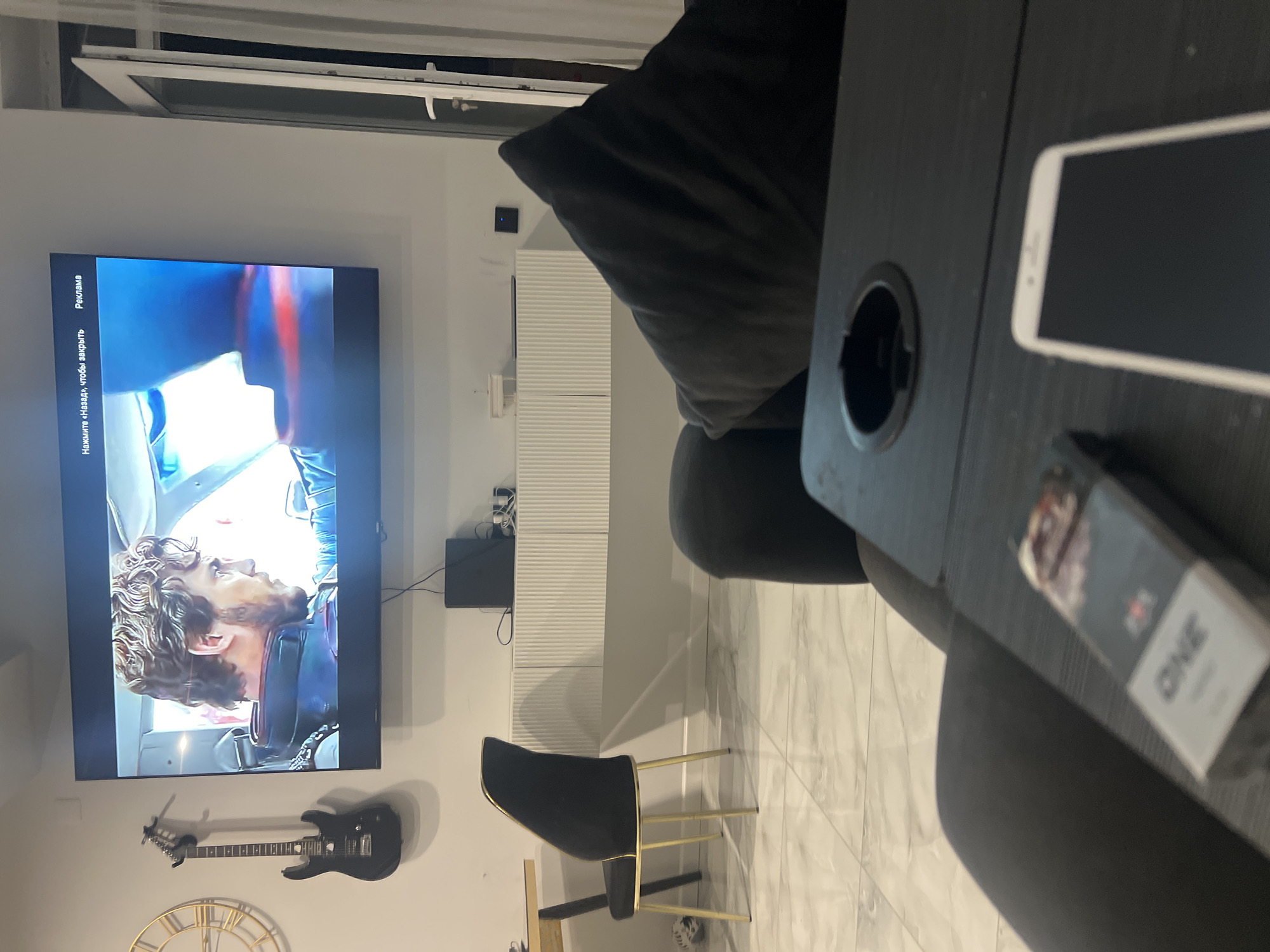 The image depicts a dimly lit living room setting. A large flat-screen television mounted on a white wall displays a movie scene featuring a close-up of a man with curly brown hair. Below the television, a white, ribbed console unit is mounted on the wall. To the left of the television, an electric guitar is hung on the wall, and a large, round clock face is visible. In the foreground, a dark gray sofa is prominent, with a dark tray table resting on its armrest. The tray table has a built-in cup holder and holds a pack of cigarettes labeled "ONE" and a white smartphone. A black dining chair with gold legs is positioned to the left of the frame, near a table. The floor is tiled with a marble pattern. The overall ambiance suggests a relaxed evening at home. The location is not specified.