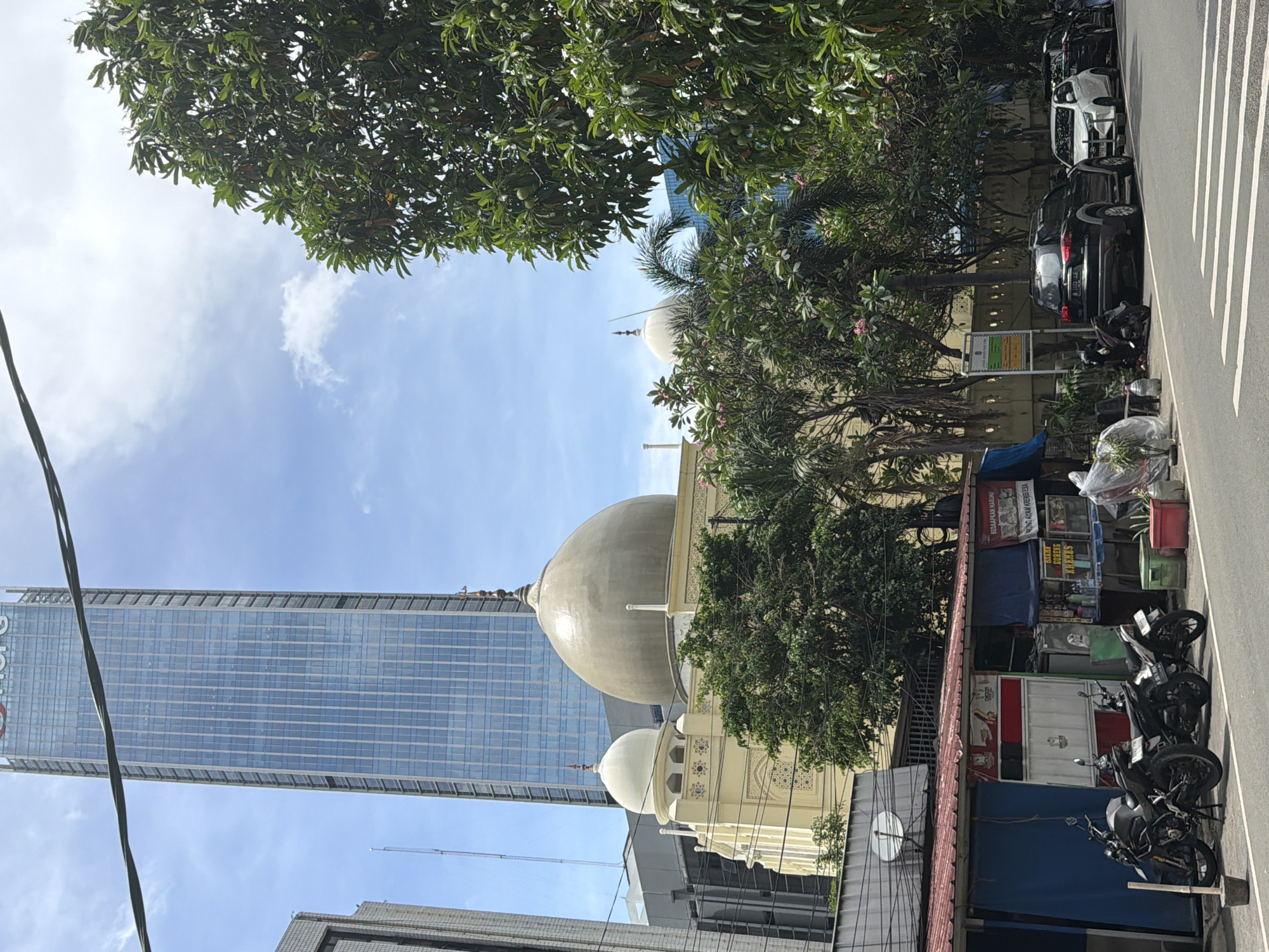 The image captures a view of South Jakarta, Indonesia, featuring a juxtaposition of modern and traditional elements. A towering glass skyscraper rises in the background against a blue sky with sparse clouds. Positioned closer is a mosque with a large central dome and smaller surrounding domes. Trees frame the mosque and skyscraper, adding a touch of greenery to the urban scene. The foreground reveals a bustling street scene with parked motorcycles, cars, and a small roadside stall. The stall appears to be selling snacks and drinks. Pedestrian crosswalk markings are visible on the street.