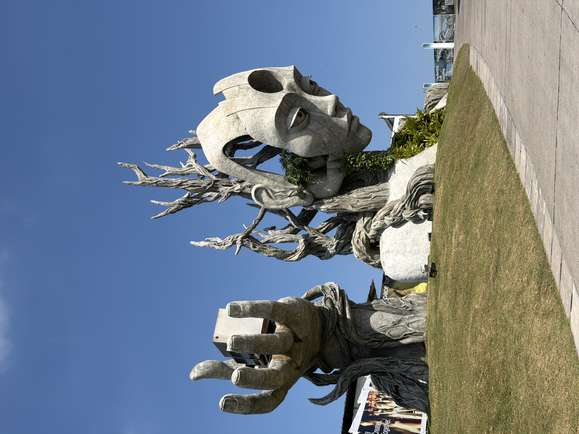 The image shows a large, abstract sculpture of a head and a hand against a clear blue sky. The sculpture is made of a stone-like material and features a stylized human face with large eyes and a neutral expression. Green vines and foliage are growing on and around parts of the sculpture. The hand is depicted in an open position, holding a rectangular, metallic object. The sculpture is situated on a grassy hill, with a paved pathway curving in front of it. In the background, there are posters and signs, suggesting a public area or attraction. One poster mentions "The Rooftop Bar for Living." The sculpture is a prominent landmark in the city of Kediri, Indonesia.
