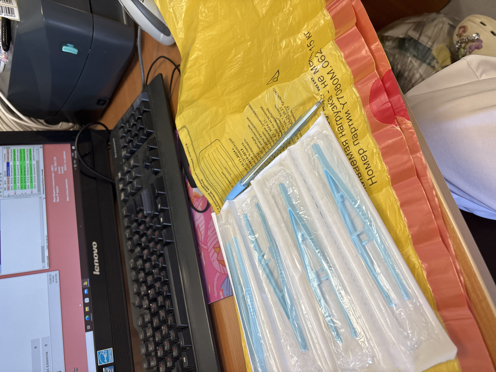 The image shows a desk with a Lenovo computer, keyboard, and a stack of sealed, blue plastic tweezers on top of several plastic bags (yellow, pink, orange). A pen rests on the tweezers. A cropped view of the computer screen shows Russian text, and the bag has Cyrillic text including "Номер партии Y7080M.062 15 кг". The text includes the name of a city, Kazan, Russia. The image does not feature Saint Petersburg or any evidence of people or actions.