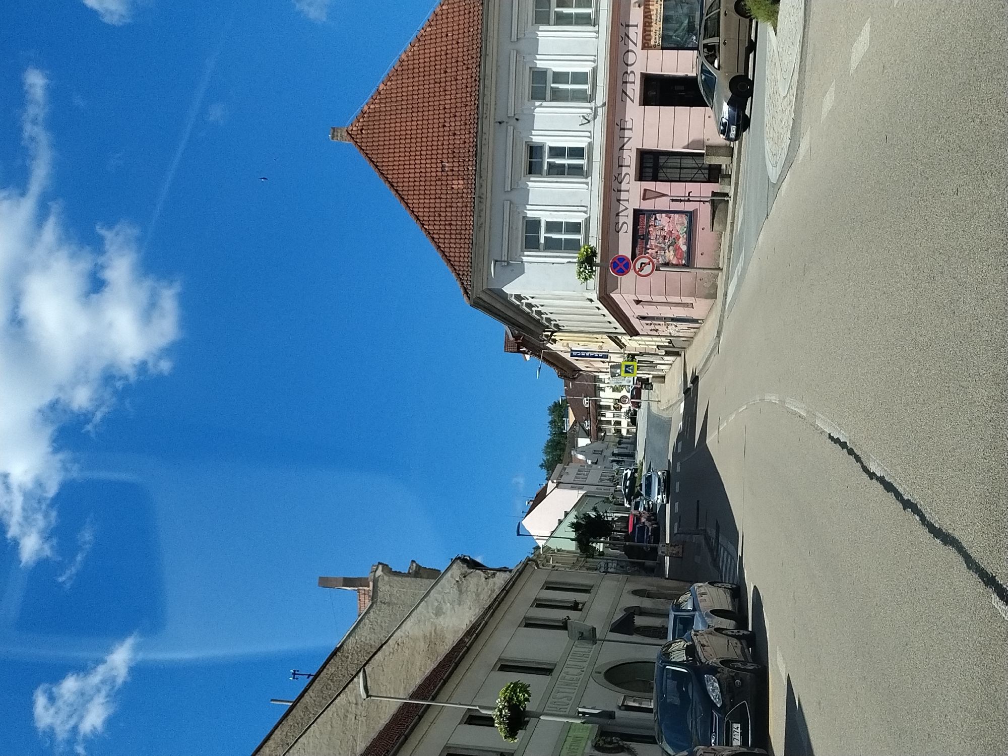 The image depicts a street scene in Volyně, Czechia, on a clear, sunny day with a bright blue sky and scattered white clouds. The perspective is from a vehicle traveling down a paved road that curves to the right.
On the left side of the street, a light-colored building with arched doorways and windows is visible, with plants in hanging baskets. Further down the street, other buildings with red-tiled roofs line the road. On the right side of the street, a pink building with white-framed windows features the signage "SMÍŠENÉ ZBOŽÍ" and "POTRAVINY". A pedestrian crossing is present in front of this building.
Several cars are parked along the street and some are in motion. A black Ford is parked on the left, and a silver station wagon is driving on the right. A few other vehicles are seen further up the road. The street is lined with trees and foliage in places. The overall impression is a typical European town street scene on a pleasant day.