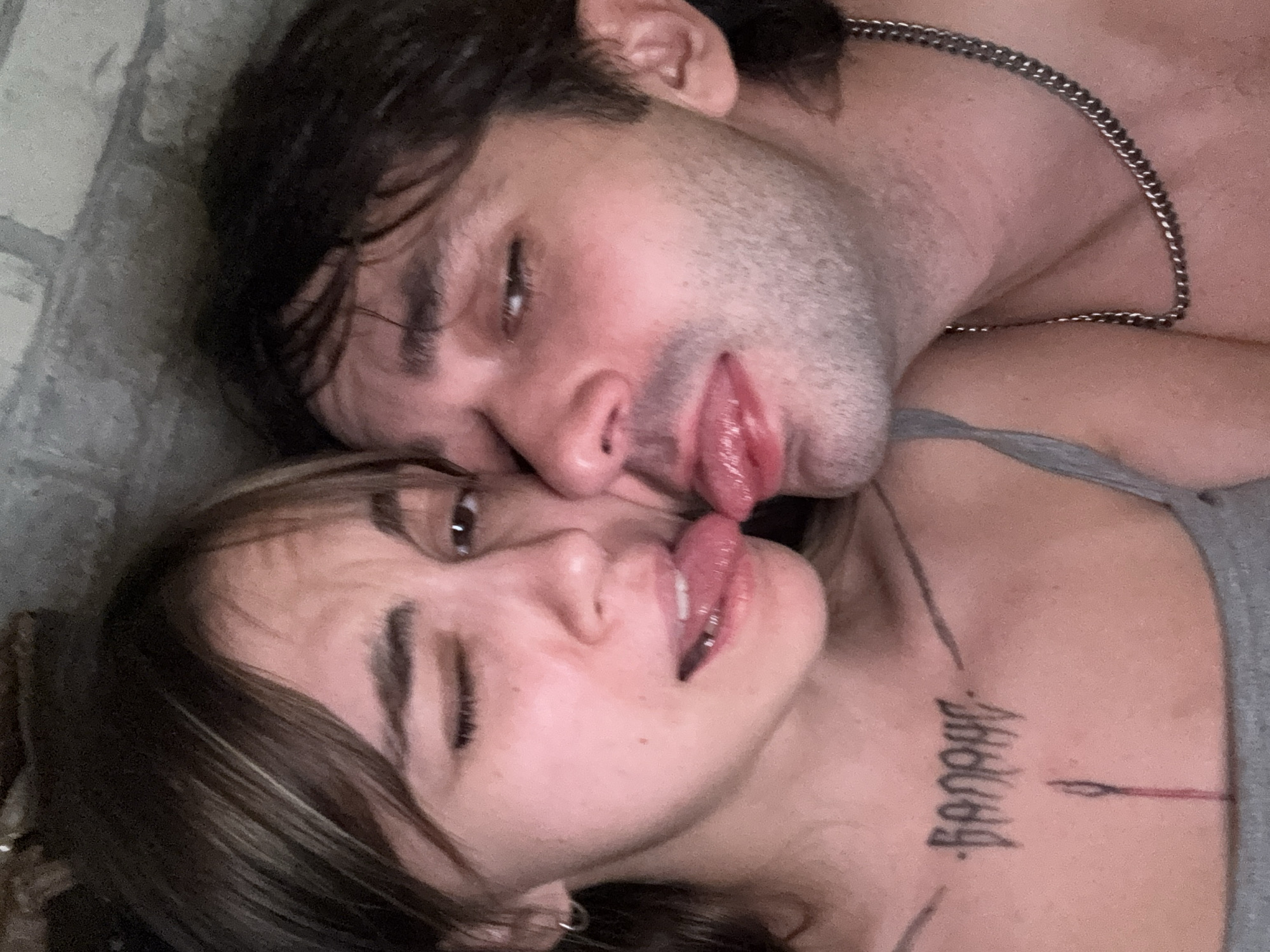 A close-up, low-angle selfie features a young woman and a young man in an intimate pose, their tongues touching. The woman, on the left, has brown hair with blonde highlights and is winking. She has a tattoo on her chest that reads "BANOHE" and appears to be a line drawing of a needle. The man, on the right, has dark brown hair and is wearing a silver chain necklace. They are positioned in front of a brick wall. There is no visible indication of the city of Moscow, Russia.