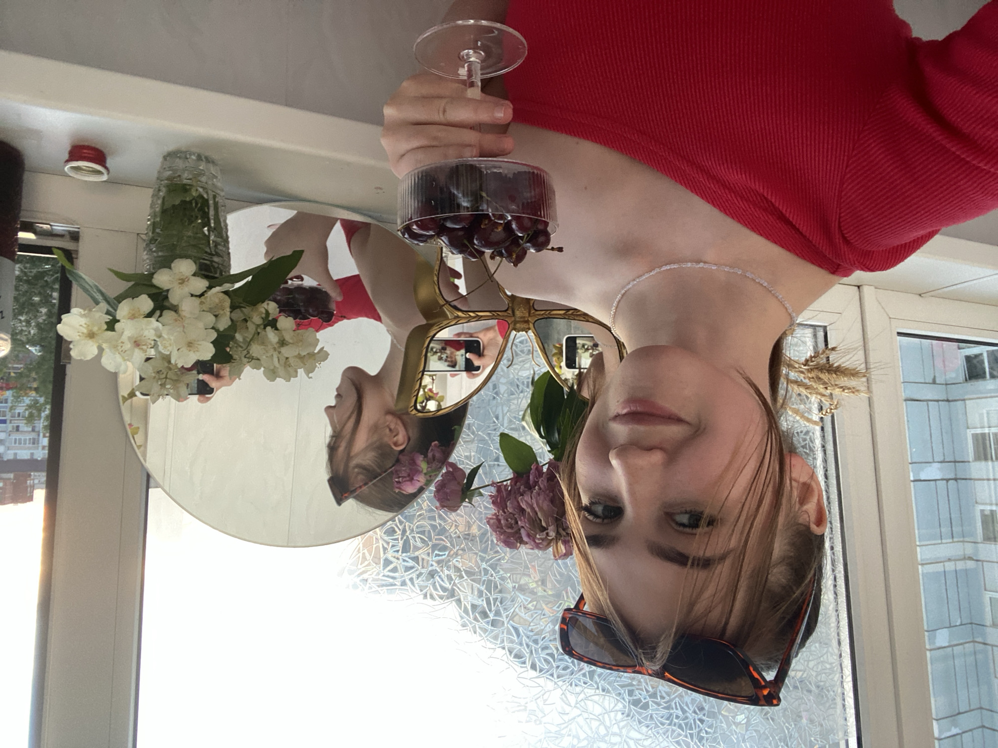 A young Caucasian woman with light brown hair, wearing a red top and sunglasses, takes a selfie in an apartment with a window and a mirror. She is holding a glass of cherries. In the background, there are buildings and trees visible through the window, indicating a city environment. The mirror reflects the woman, a vase of white flowers, and more buildings. The scene appears to be set during the daytime. It is not possible to determine the specific location in Chaikovsky, Russia from the image.