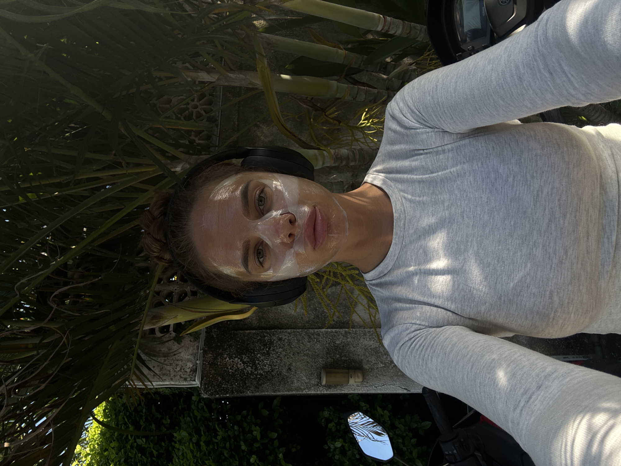 A woman with a white facial mask and black headphones is seated on a motorbike. She wears a light grey long-sleeved shirt. Palm trees and other foliage are visible in the background, as well as a concrete wall or structure. The motorcycle has a digital display and side mirrors reflecting the surroundings. The image was taken outdoors in North Kuta, Indonesia.