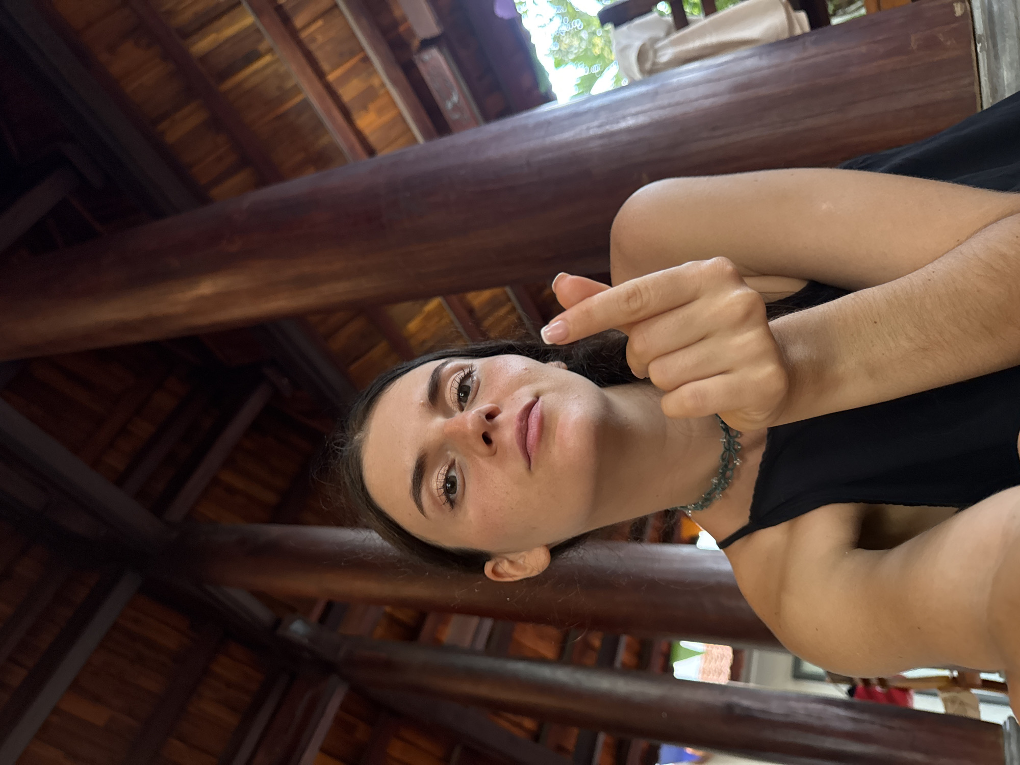 This image captures a young woman in what appears to be a shaded, semi-outdoor setting within Mũi Né, Vietnam. The photograph is tilted, presenting the subject and background at an angle.
The main subject is a young woman, positioned looking up and slightly to the side towards the camera. She has dark hair, light-colored eyes, and a neutral expression, enhanced by what appears to be subtle makeup. She is wearing a simple black top or dress, and a distinctive beaded or woven green necklace is visible around her neck. Her right hand is raised, making a "finger heart" gesture, commonly used to express affection or friendliness.
The background is dominated by the intricate wooden structure of a ceiling, featuring dark wooden beams and planks. Beyond this structure, bright daylight filters through, revealing lush green foliage, suggesting an outdoor, natural environment. The warm lighting implies it is daytime. The architectural style, combined with the visible greenery and the Mũi Né context, points to a tropical resort, traditional building, or open-air cafe setting. No visible text is present in the image.