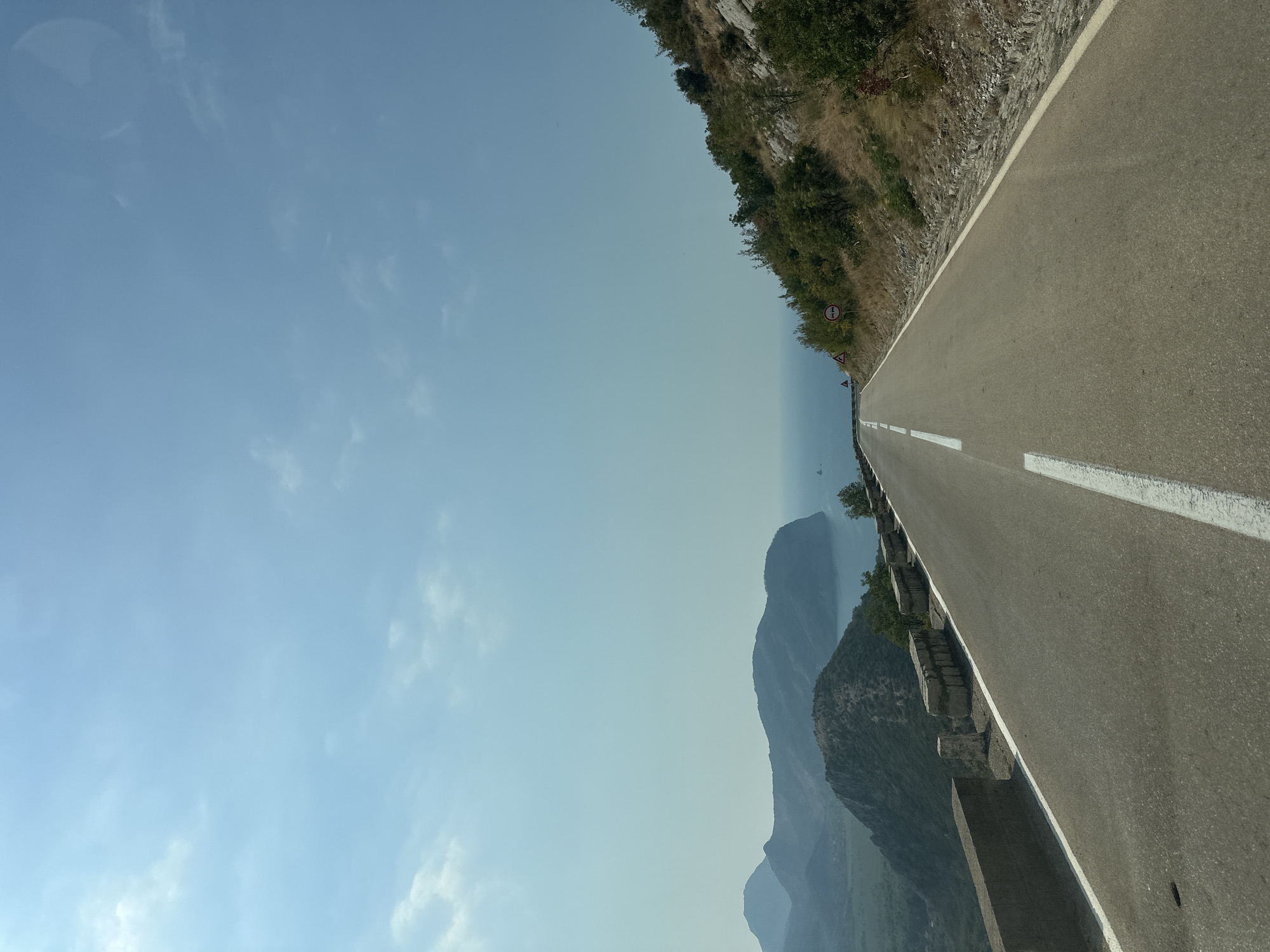 The image depicts a scenic view of a winding road ascending a mountainside, overlooking a body of water and distant hills. The road is paved, with a white dashed line marking the center and a solid white line along the right edge. Guardrails made of stone blocks are visible along the left side of the road. The surrounding vegetation consists of dry grass, shrubs, and scattered trees, indicative of a Mediterranean climate. The sky is clear and blue, with a few wispy clouds. Traffic signs, including a triangular warning sign with a symbol and a circular sign with a red border and a symbol, are present on the right side of the road. The overall scene suggests a journey through a mountainous coastal region. This location is consistent with the descriptions of roads and landscapes found in and around Novoselje, Montenegro, a settlement known for its proximity to the Adriatic Sea and its mountainous terrain.