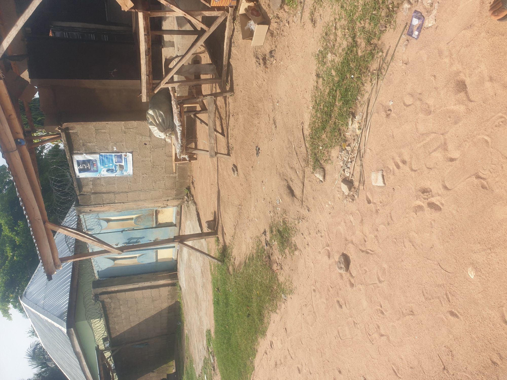 The image depicts a scene in Jauro Yinu, Nigeria, featuring a dirt road leading towards a compound. To the left, a building with a corrugated metal roof and light green walls is visible, bordered by a brick wall topped with barbed wire. A blue and yellow metal gate is set within this wall. To the right, a rustic wooden structure with a thatched or corrugated roof stands adjacent to a brick building. A large, light-colored sack is placed on a wooden bench in front of this building, next to a poster advertising laundry services. The ground is covered in sandy soil with scattered rocks and patches of grass. Footprints are visible in the sand, indicating recent activity. A discarded cardboard box with some items inside is located on the ground to the right of the wooden structure. The environment appears to be a semi-urban or rural setting.