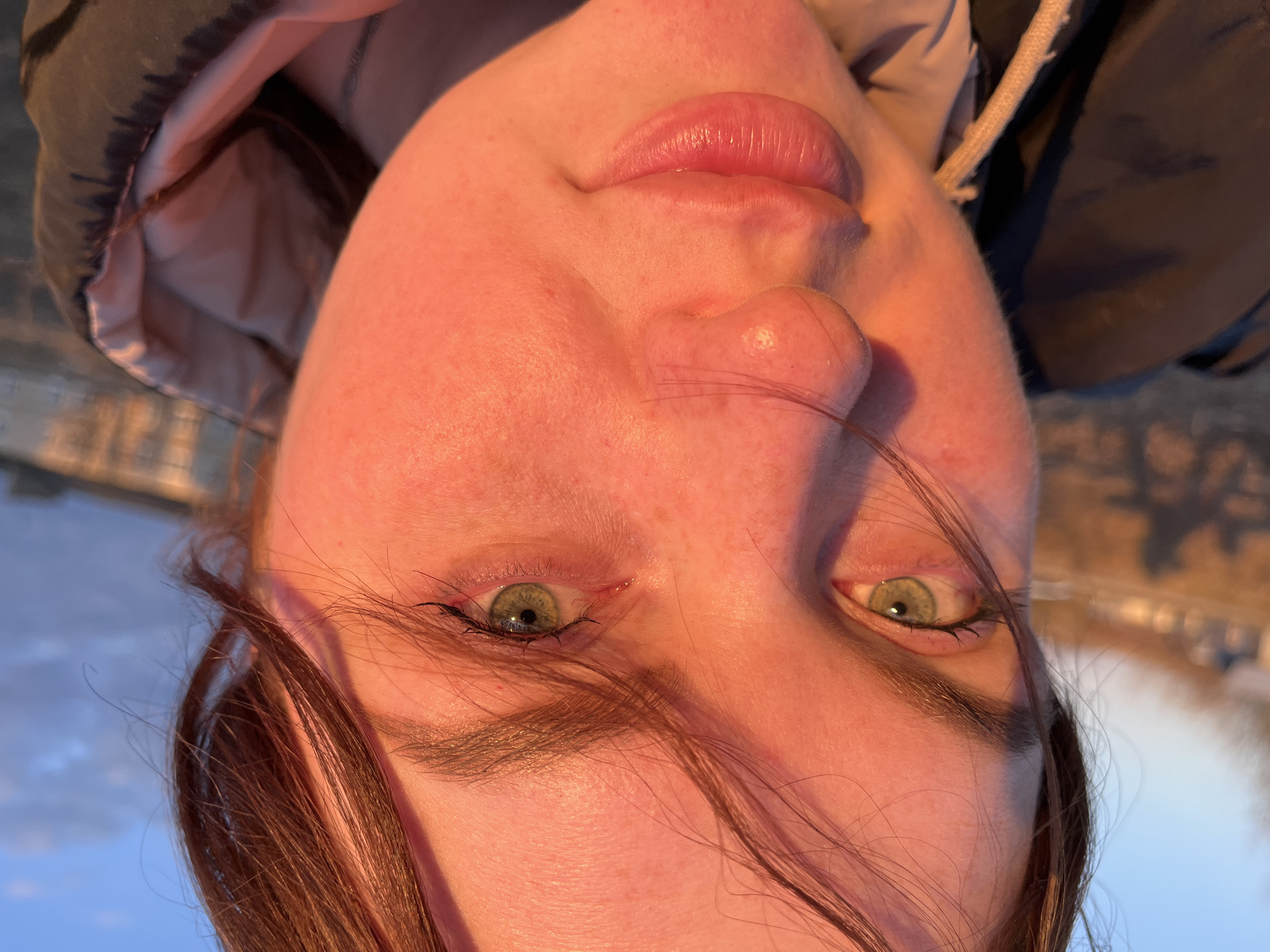 A close-up, inverted photograph depicts a person with fair skin, hazel-green eyes, and light brown hair. The subject is wearing a dark-colored hooded jacket. The background shows a blurred outdoor scene with light blue and brown hues, possibly a river or field during sunset. The location may be Павловка, Russia.