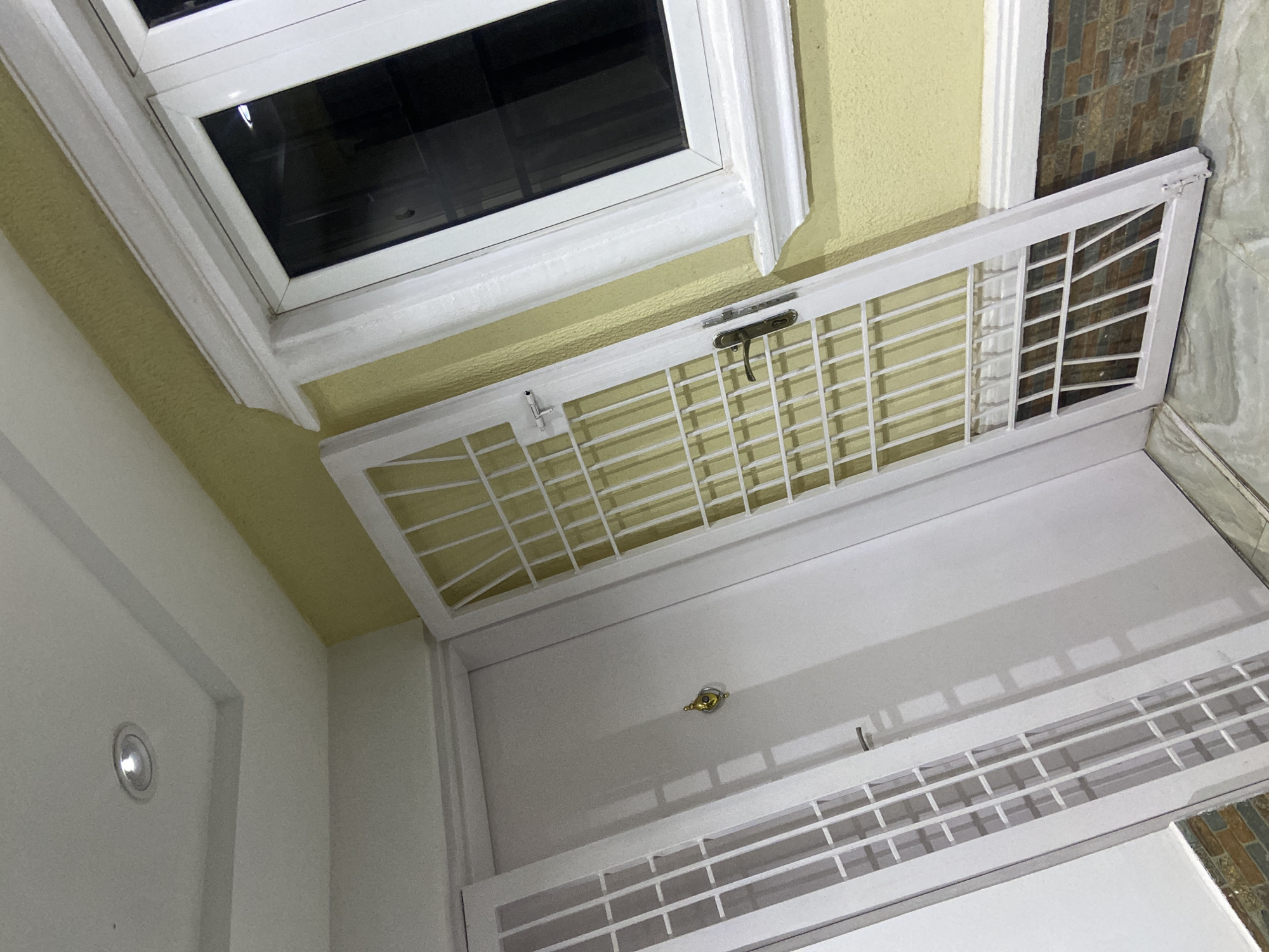 The image captures a close-up, angled view of an exterior doorway with a white metal security gate. The doorway appears to be part of a building with yellow-painted walls and white trim around a window on the right. The floor is tiled with a marble-like pattern. A recessed light fixture is visible in the white ceiling. The security gate is open and leans against the wall, revealing a white door behind it. The door has a circular, gold-toned door handle. The scene suggests a residential or commercial property. The location is identified as Gura Top, Nigeria.