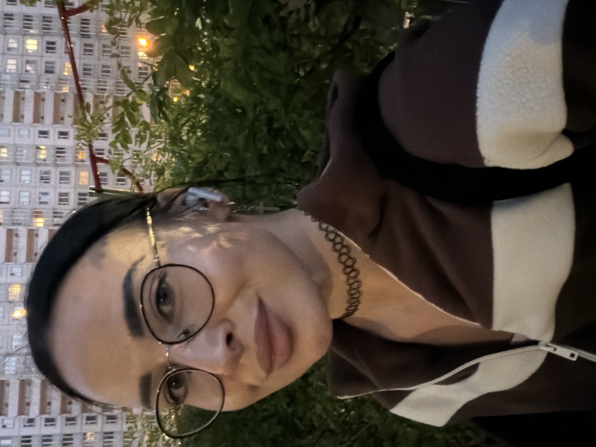 An evening selfie captures a person in Saint Petersburg, Russia, with an urban background. The subject, a person with dark hair and glasses, looks directly at the camera with a neutral to slight smile. They are wearing a dark brown hooded jacket or sweatshirt featuring a broad white stripe on the side and arm, with a visible zipper detail. A black choker necklace is worn around their neck. The background features a large, light-colored residential building with numerous windows, many of which are illuminated, suggesting it is nighttime or late evening. Dark green foliage, likely from trees or bushes, partially obscures the building on the right side of the frame. A reddish structural element is also visible in the background, possibly part of a building or a nearby object. No discernible text is visible in the image.