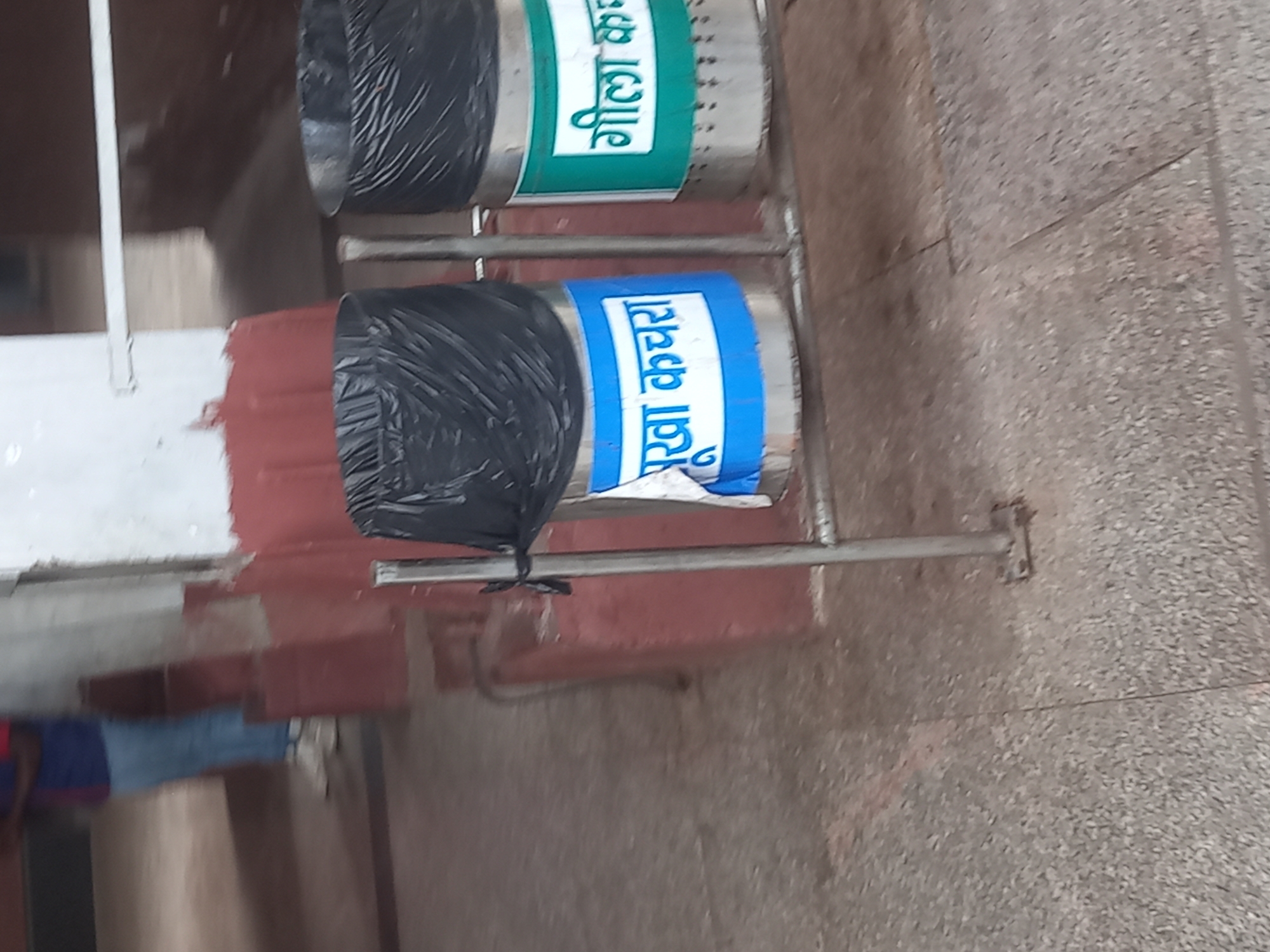 Two metal trash cans are stacked on a metal frame. The top trash can is labeled "Geela Kachra" (wet waste) with green text. The lower trash can is labeled "Sukha Kachra" (dry waste) with blue text. Both trash cans are covered with black plastic bags. The background consists of a light-colored wall and floor. The scene appears to be in an outdoor public area in Jhansi, India.