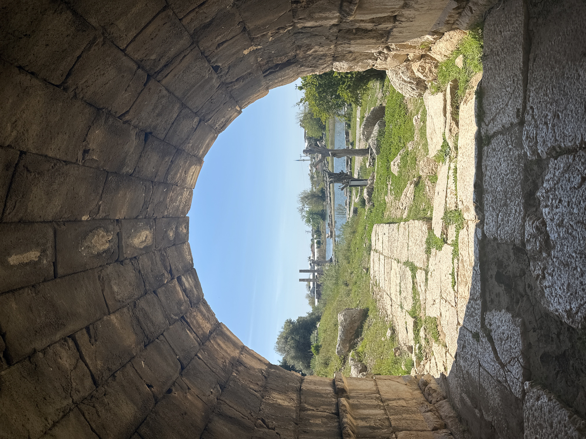 View through an ancient stone arch in Seydikemer, Turkey. The arch is constructed of large, rough-hewn blocks. Outside the arch, a stone path leads toward a body of water. Visible along the path and beyond are remnants of ancient structures, including columns and architectural fragments, interspersed with grass and foliage. The sky is clear and blue. Two dead tree sculptures are erected in the midground of the frame.