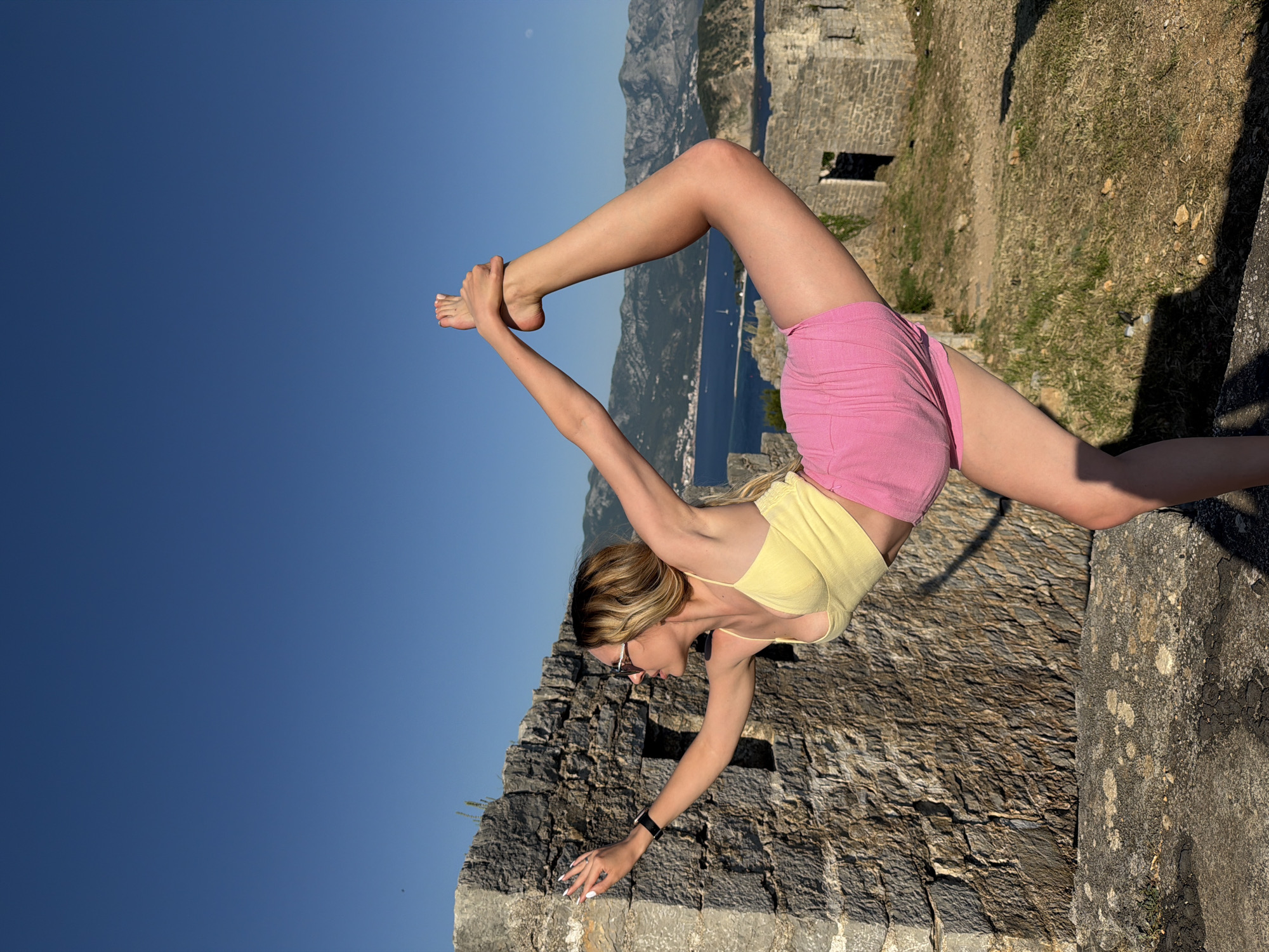 A woman is performing an extreme yoga pose on a stone wall in Budva, Montenegro. She is wearing a yellow top and pink shorts and has sunglasses on. Her left leg is on the wall, and her right leg is extended upward, grasping her foot with her right hand. Her left hand is touching the stone wall for balance. The background shows a view of the sea, mountains, and a stone building in the distance. The sky is clear and blue.