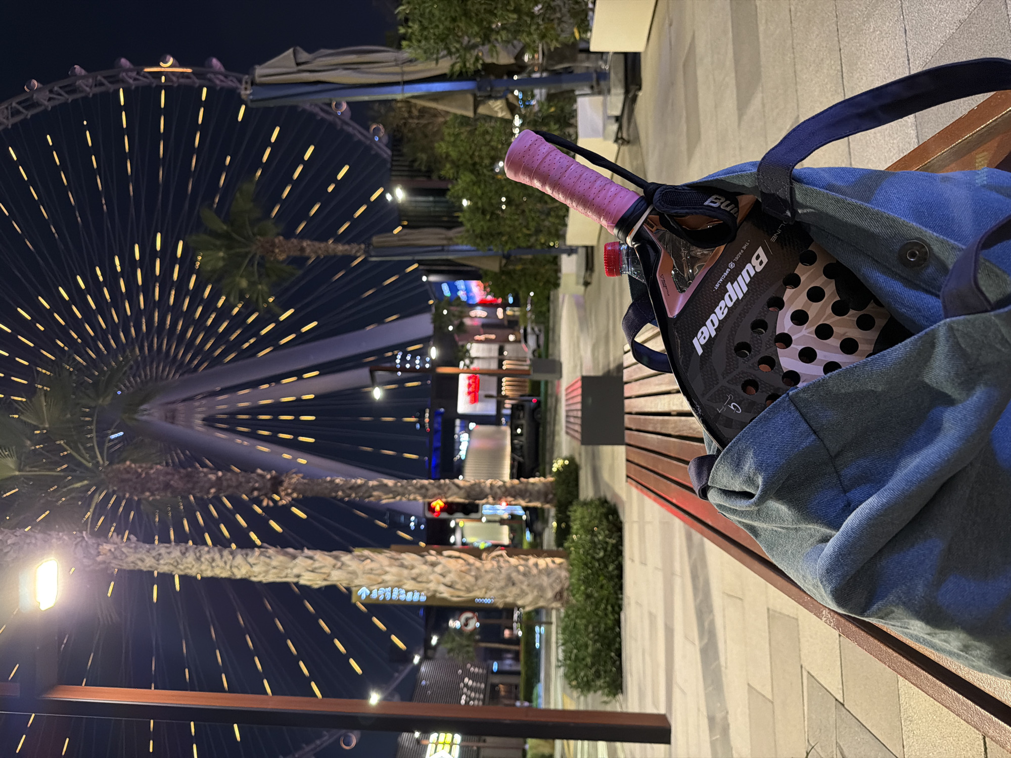 The image captures an urban night scene in Dubai, United Arab Emirates, featuring a padel racket placed within a bag on a wooden bench in the foreground. The time is clearly night, evidenced by the dark sky and numerous artificial lights illuminating the area.
In the immediate foreground, a casual, blue denim-like bag rests on a slatted wooden bench. Peeking out from the bag is the top portion of a padel racket, featuring a striking pink grip. The racket itself is black with pink accents, and its face displays multiple circular holes. The brand "Bullpadel" is clearly visible on the racket, along with the slogan "• THE PADEL SPECIALIST •". A red cap of a water bottle is also partially visible near the racket's grip.
The background is dominated by the illuminated structure of a large Ferris wheel, likely Ain Dubai, with countless small yellow lights radiating from its spokes. Several tall palm trees are also visible, some partially obscuring the Ferris wheel, and their fronds are faintly lit by surrounding light sources. Beyond the Ferris wheel and trees, various buildings and structures are lit with colorful lights, contributing to a vibrant city atmosphere. Faint text and directional arrows can be seen on distant signboards, though most are indistinct due to distance and focus. One visible sequence appears to be "↑ 83 83 83" on a blurred sign.
No people are visible in the photograph, creating a tranquil urban snapshot. The presence of the padel racket suggests a pause in or anticipation of a sports activity within a bustling city setting. The weather appears clear and calm, suitable for an evening stroll or outdoor activity.