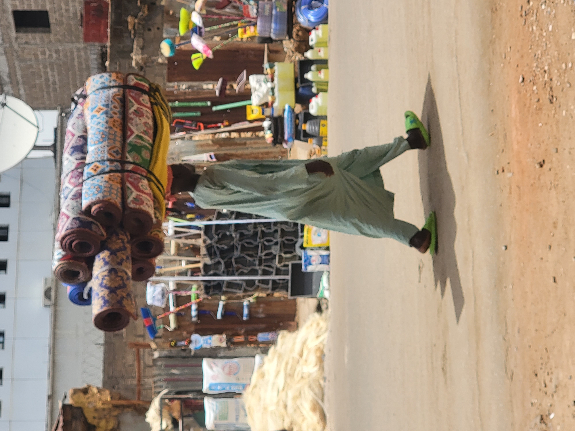 The image depicts a street or market scene, oriented incorrectly and showing a man walking under bright daylight. The man, dressed in a light green or grey traditional garment and bright green flip-flops, carries a tall, bundled stack of colorful, patterned rolled mats or rugs, secured with straps. His short, distinct shadow indicates a high sun, suggesting it is mid-day.
Behind the man, an open-front shop or stall is visible, displaying various goods. These include numerous yellow and blue plastic containers, possibly jerricans or bottles, alongside cleaning supplies such as brooms, mops, and brushes. A coiled blue hose is also part of the displayed merchandise. On the ground near the stall, white bags are piled, with one clearly showing the text "ULTIMATE". Next to these bags are piles of light-colored fibers or string.
To the man's left, a building with dark, rectangular windows is partially seen, featuring a white satellite dish mounted on its exterior. The ground is a mix of light-colored pavement and a dusty, reddish-brown area with small stones. The scene captures a moment of daily activity and commerce in an urban or town setting, consistent with a market in Maiburiji, Nigeria. No specific emotions are discernible from the man's posture or partially visible face.