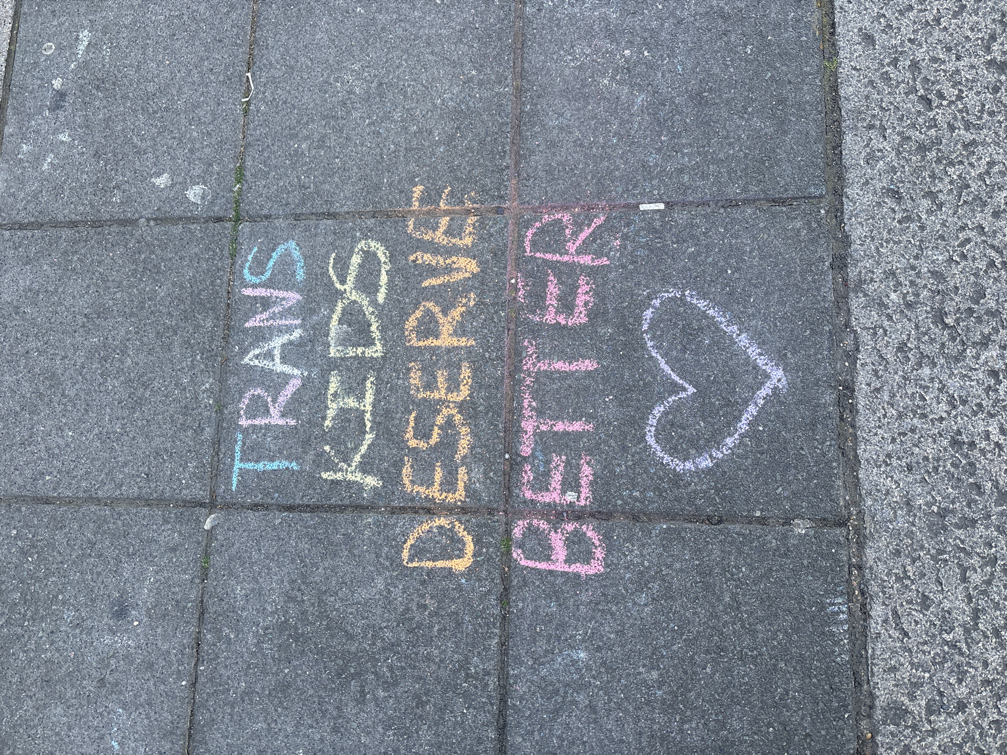 The image shows a section of a gray paved sidewalk in a city. The message "TRANS DESERVE BETTER" is written in colorful chalk on four adjacent paving stones. The letters are vertically oriented and stacked on top of one another. The colors of the chalk used include blue, pink, yellow, and orange. A white chalk heart is drawn on the paving stone to the right of the word "BETTER". A small piece of litter is visible on the upper right side of the paving stone with the word "TRANS". The image does not reveal any explicit details confirming that the location is the city of London, United Kingdom.