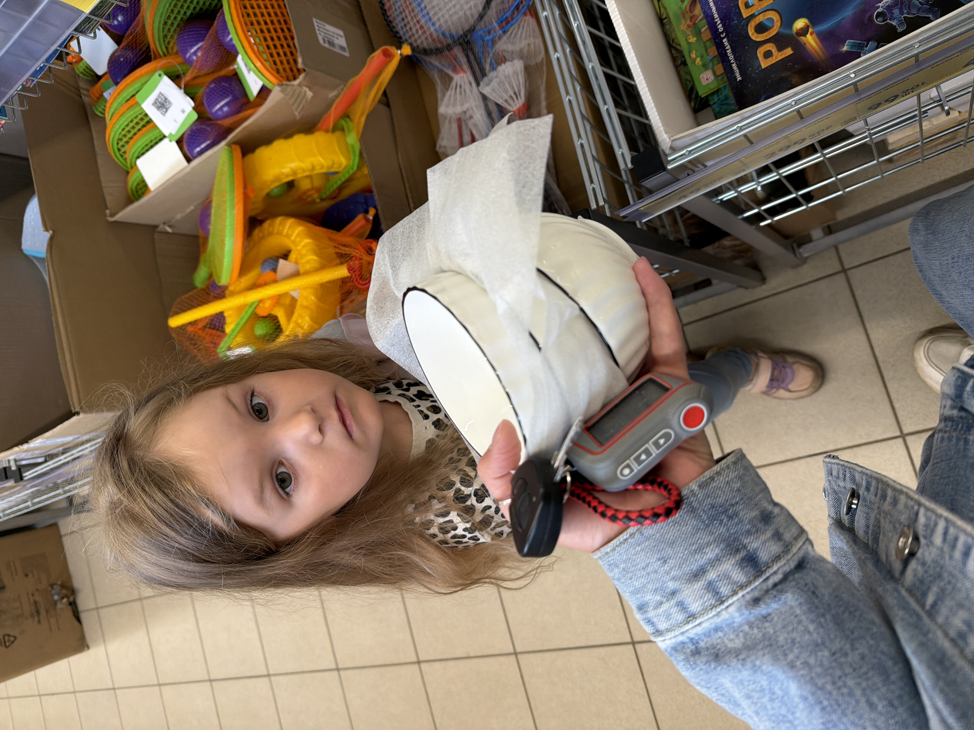 The image captures a candid moment in a store aisle, viewed from a slightly upward angle. A young child, likely a girl with light brown hair and a calm expression, looks directly at the camera. She is wearing a leopard print top. In the foreground, an adult's hand, presumably holding the camera, displays a white ceramic bowl or dish wrapped in protective foam and a set of car keys with a gray remote fob attached to a red and black braided strap. The adult wears a denim jacket, and their white shoe is visible at the bottom right. The child's purple-brown shoes are also visible on the tiled floor.
The setting is a retail environment, characterized by light-colored square floor tiles and metal shelving units. To the left, a cardboard box overflows with colorful children's toys, including orange and green mesh rackets and yellow plastic beach toys, some bearing green tags with QR codes. Above these toys, badminton rackets are visible on a rack. To the right, on another shelf, a book or board game box with an astronaut and space theme is displayed. Visible Cyrillic text on this box includes "РОБ" (likely part of "РОБОТЫ" or "РОБОТ") and "Энциклопедия с объемными" (Encyclopedia with volumetric/3D). Price tags below the shelf show numbers like "299", "189", and "99", suggesting prices in rubles. The scene depicts a casual shopping trip.