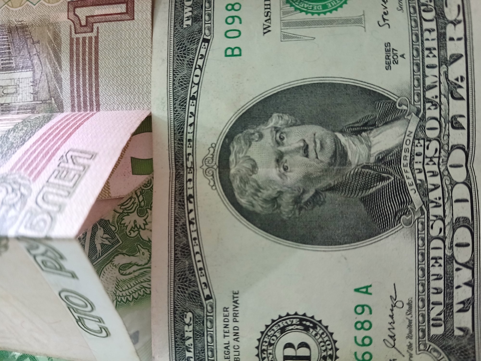The image shows a close-up of a two-dollar bill with the portrait of Thomas Jefferson. Partially visible alongside the two-dollar bill is what appears to be a Russian ruble. The ruble is folded over, partially obscuring the edges of the two-dollar bill.
