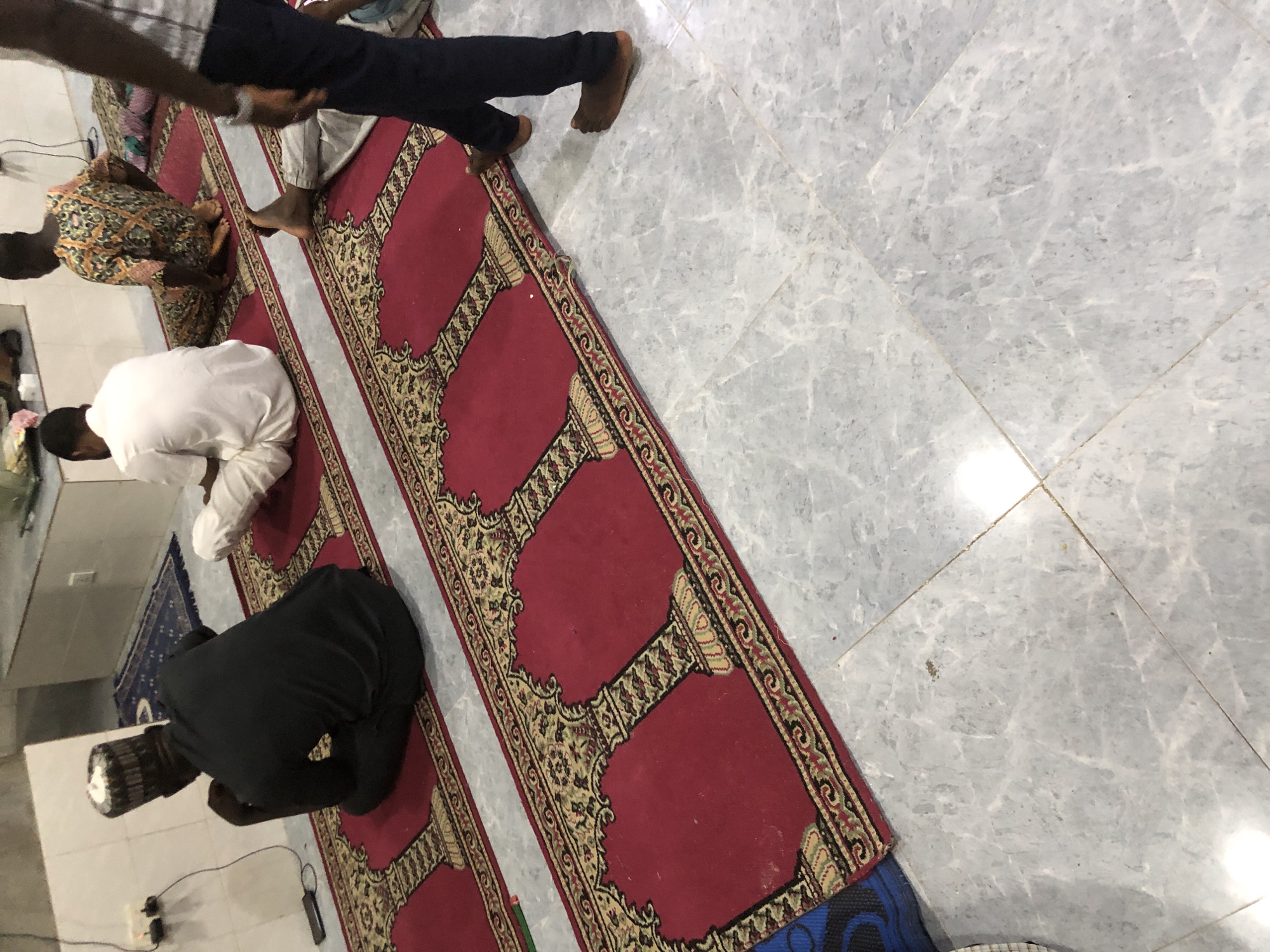 The image depicts a group of men seated and kneeling on prayer mats in what appears to be a mosque or prayer hall. The floor is tiled with a light gray marble pattern. A red prayer rug with intricate gold and black patterns runs horizontally across the frame, featuring designs that resemble arches.
On the left, a man wearing a black garment and a patterned kufi hat is seated on the rug, facing away from the camera. To his right, a man in a white traditional outfit is kneeling. Further to the right, another man in a colorful, patterned garment is also kneeling. In the foreground, on the right side of the image, the lower legs and feet of a person wearing dark blue trousers are visible, with bare feet on the tiled floor. Another person's legs and feet, in light-colored trousers and bare feet, are visible lying down on a prayer mat. The lighting in the room is somewhat dim, with some overhead light sources casting reflections on the floor. Electrical outlets and cords are visible on the wall in the background on the left.
This scene is described as being located in Ado, Nigeria, suggesting a place of worship within that city.