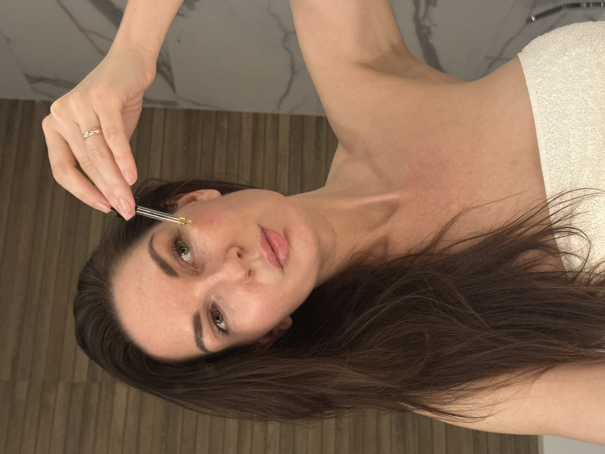 A woman with brown hair is applying oil to her face using a glass dropper. She wears a wedding ring. A white towel is draped over her shoulder. The background consists of wood paneling and a marble-patterned wall. There is no information to confirm that this image was taken in the city of Chaikovsky, Russia.