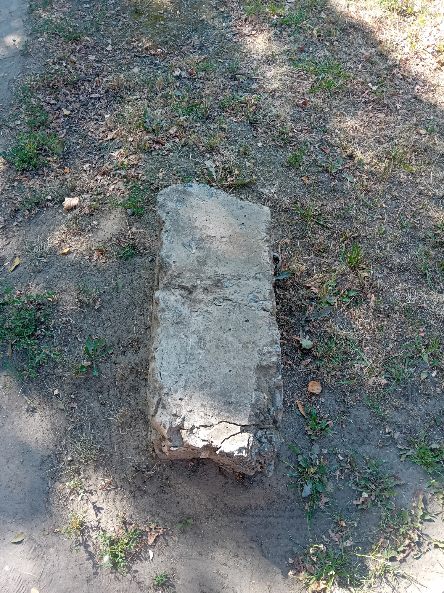 The image shows a concrete block lying on the ground in a park or natural area in Chuhuiv, Ukraine. The block is rectangular and appears to be weathered and damaged, with cracks and rough edges. It is surrounded by dry grass, scattered leaves, and patches of dirt. Sunlight casts shadows on the ground and the block, indicating it is daytime. There are no people or animals visible in the image. The scene suggests a public outdoor space, possibly a neglected or undeveloped area within the city.