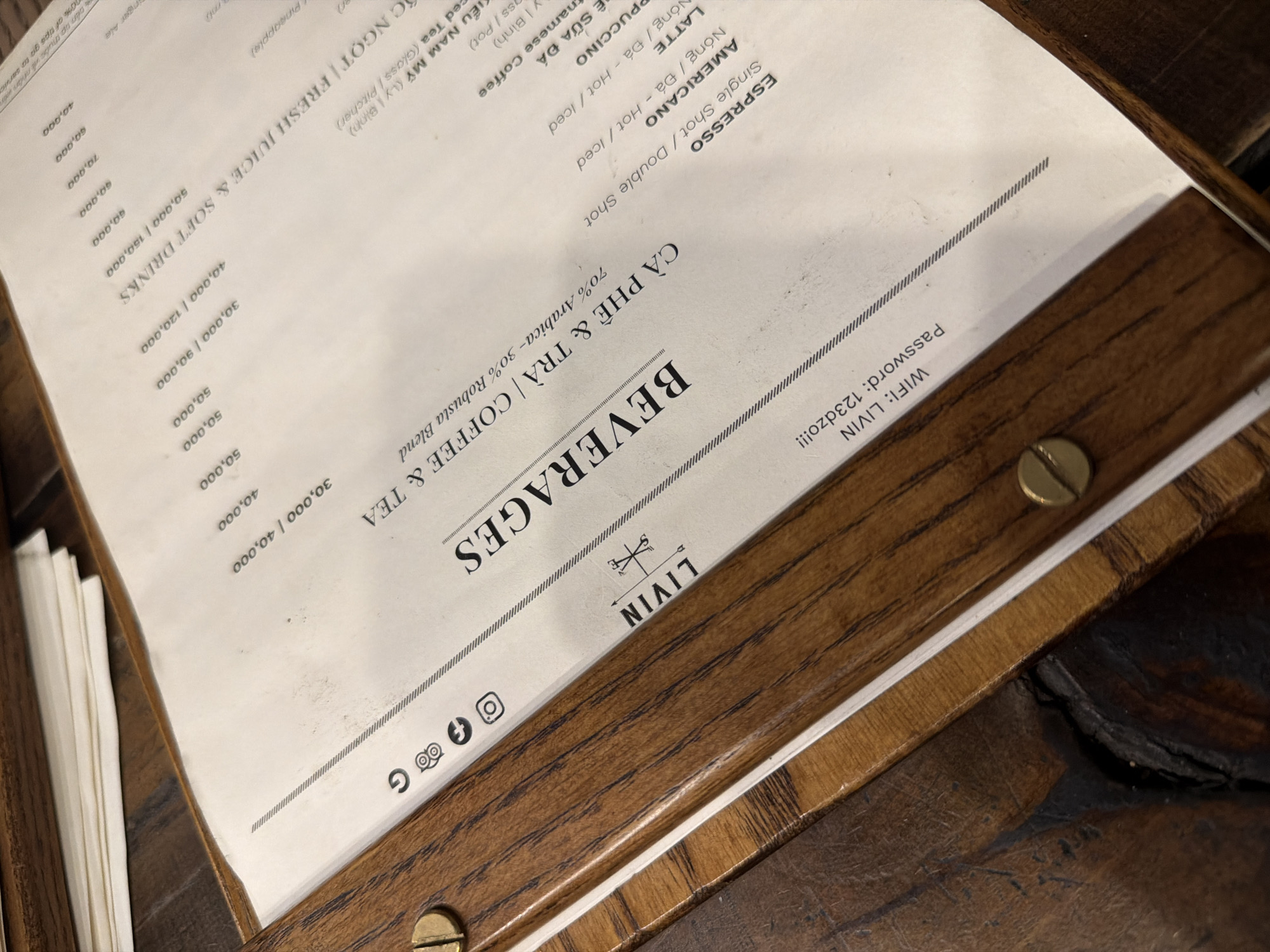 A slightly high-angle, close-up shot shows a wooden-bound menu, presumably from LIVIN in Nha Trang, Vietnam. The menu lists beverages, including coffee and tea, and shows the Wi-Fi password, "123dzo!!!". The menu cover is light cream colored with brown text. The wood binding has brass bolts securing it. The table that the menu is sitting on is stained dark brown. Other menus can be seen in the lower-left, behind the top menu.