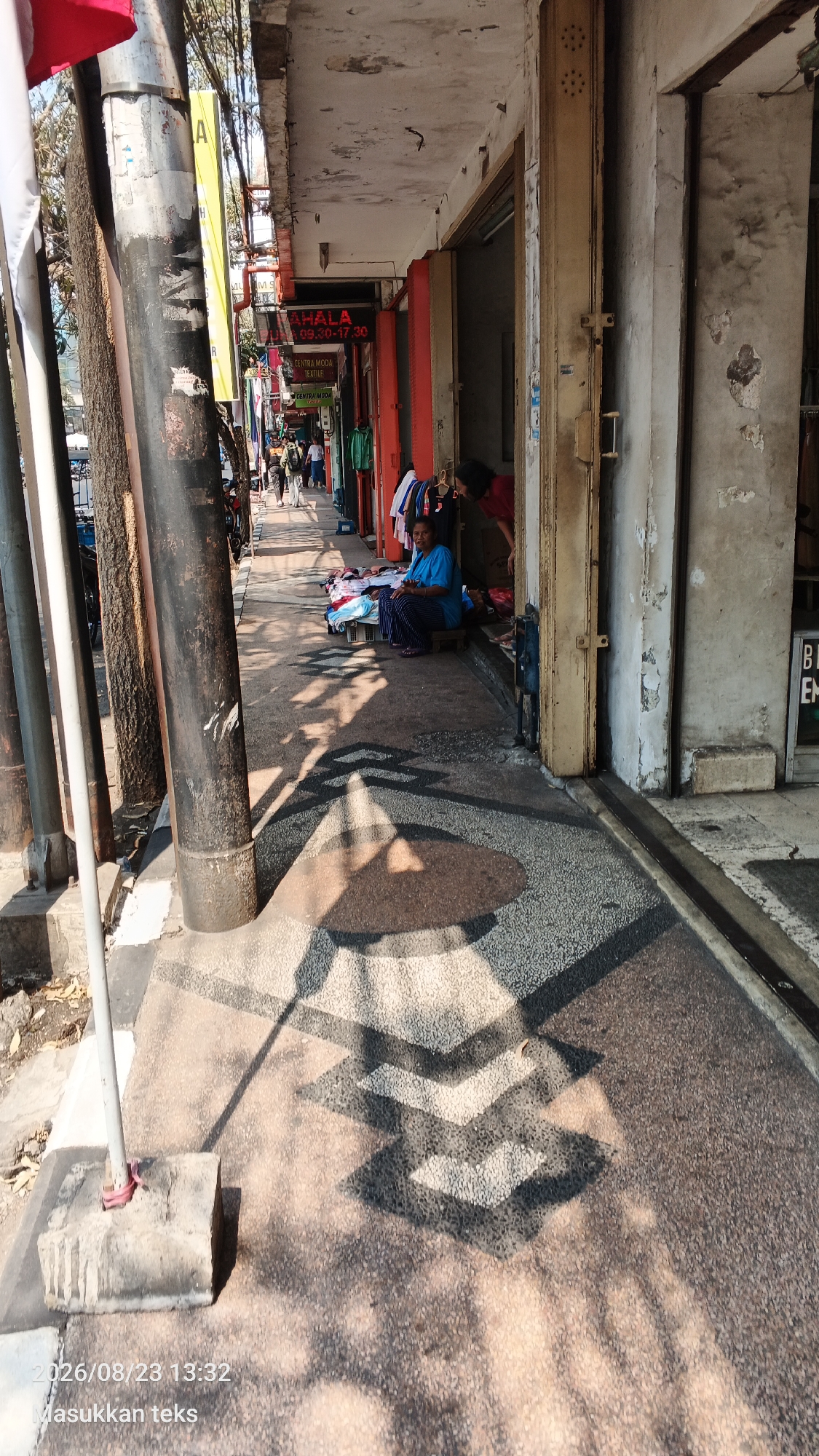 This is a daytime, eye-level, outdoor shot on a sidewalk in the city of Malang, Indonesia, showing a woman sitting on a mat surrounded by clothing. She is wearing a blue top and dark patterned pants. Behind her, a second person, wearing a red shirt, is partially visible near a shop entrance. To the left, the sidewalk is lined with utility poles and trees, with more shops and people visible further down the street. Sunlight casts strong geometric shadows on the textured sidewalk. The scene appears to be in front of a commercial establishment.