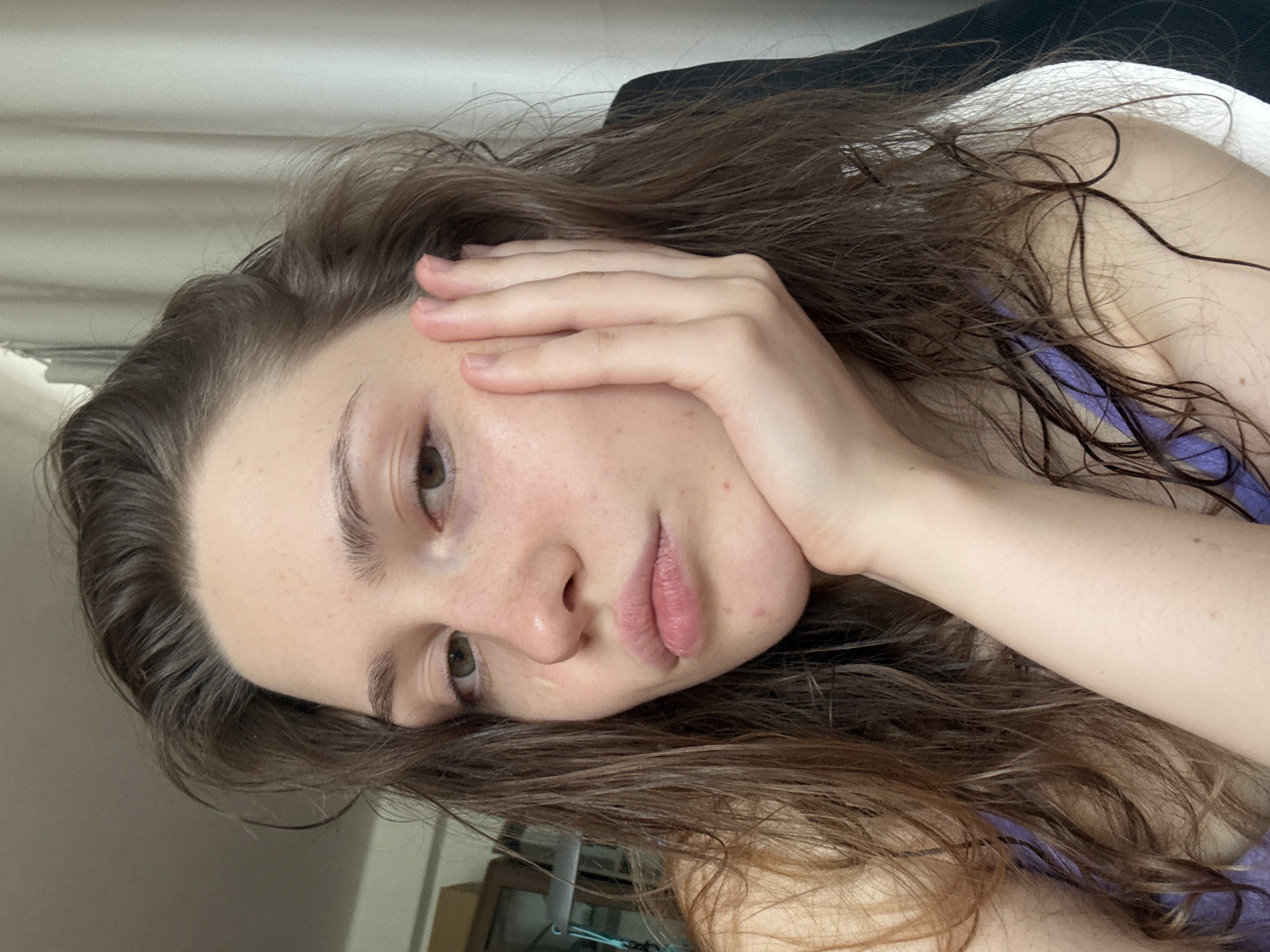 A close-up portrait shows a woman with light skin and long, wavy brown hair that appears damp. Her face is resting on her hand, with her fingers near her forehead. She has light-colored eyes and her lips are closed. She is wearing a purple tank top. There are no indications of the location being Saint Petersburg, Russia. The background is out of focus, with a hint of furniture.