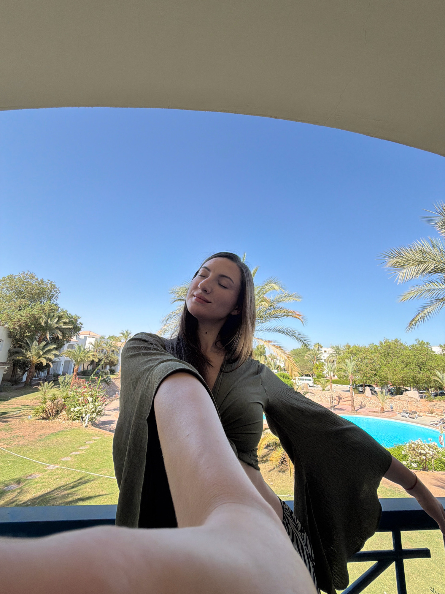 A young woman with closed eyes and a serene expression is captured in a selfie taken on a balcony overlooking a resort in Sharm El Sheikh, Egypt. She is wearing a flowing olive-green top with wide, bell-shaped sleeves and a patterned skirt. Her right arm is extended towards the camera, and her left arm is resting on the balcony railing. The background reveals a lush green lawn, palm trees, resort buildings, and a bright blue swimming pool. The sky is a clear, vibrant blue, indicating a sunny day. The overall scene suggests a peaceful vacation moment.
