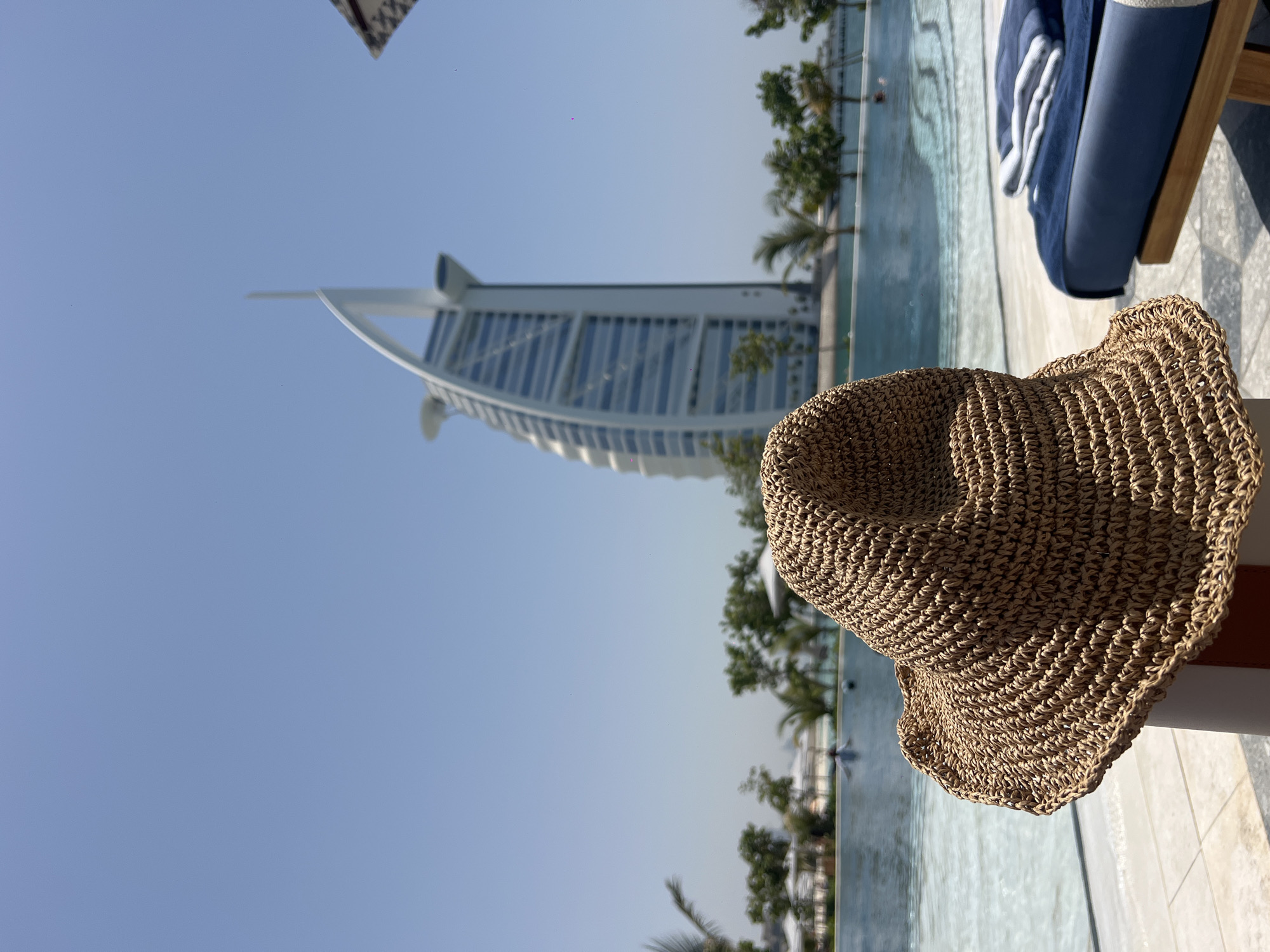 The image features a woven sunhat resting on a surface near a pool. In the background, the Burj Al Arab, a prominent luxury hotel in Dubai, is visible. Palm trees are scattered around the pool area, suggesting a tropical or resort-like setting. The sky is clear and blue, indicating sunny weather. The pool appears to be surrounded by lounge chairs, one of which is partially visible in the foreground. The overall scene suggests a vacation or leisure setting at a high-end destination in Dubai.