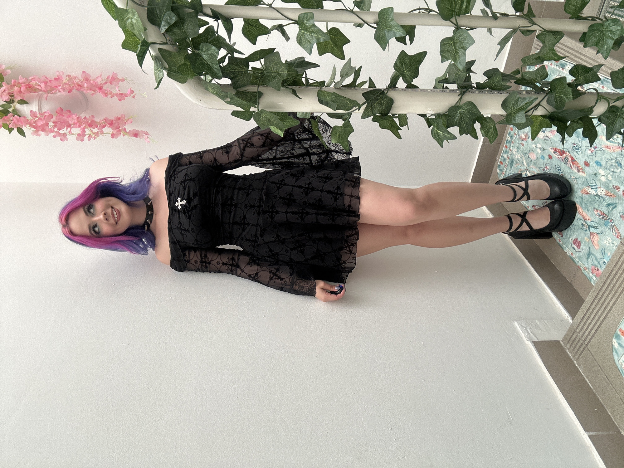 A full-body portrait, rotated 90 degrees clockwise, captures a young woman standing and posing indoors in the City of Zagreb, Croatia. She appears to be in her late teens or early twenties with light skin and a gentle smile, looking directly towards the camera. Her long hair is distinctly dyed in vibrant shades of purple and pink. She is dressed in a black, long-sleeved dress made of lace-patterned fabric, with noticeable bell sleeves. A silver-colored cross pendant adorns her chest, and she wears a black studded choker around her neck. Her feet are clad in black flat shoes with cross-straps over the instep. A ring, possibly with a blue detail, is visible on her right pinky finger.
The background consists of a bright, light-colored (likely white) wall and floor. Decorative elements are prominent: artificial green ivy is draped along a white railing or pipe positioned above her head (when corrected for rotation). Delicate pink artificial flowers, resembling wisteria or cherry blossoms, hang from a white pot or fixture further up the wall. To her left (when corrected), a patterned rug or mat is visible on what would be the floor, featuring light blue and green hues with colorful insect motifs such as butterflies, beetles, and dragonflies in red, orange, and white. Adjacent to the rug, a grey tiled step or platform is seen. The lighting is bright and even, suggesting a well-lit indoor environment. The woman's pose is relaxed, and her facial expression is pleasant. There is no visible text in the image.