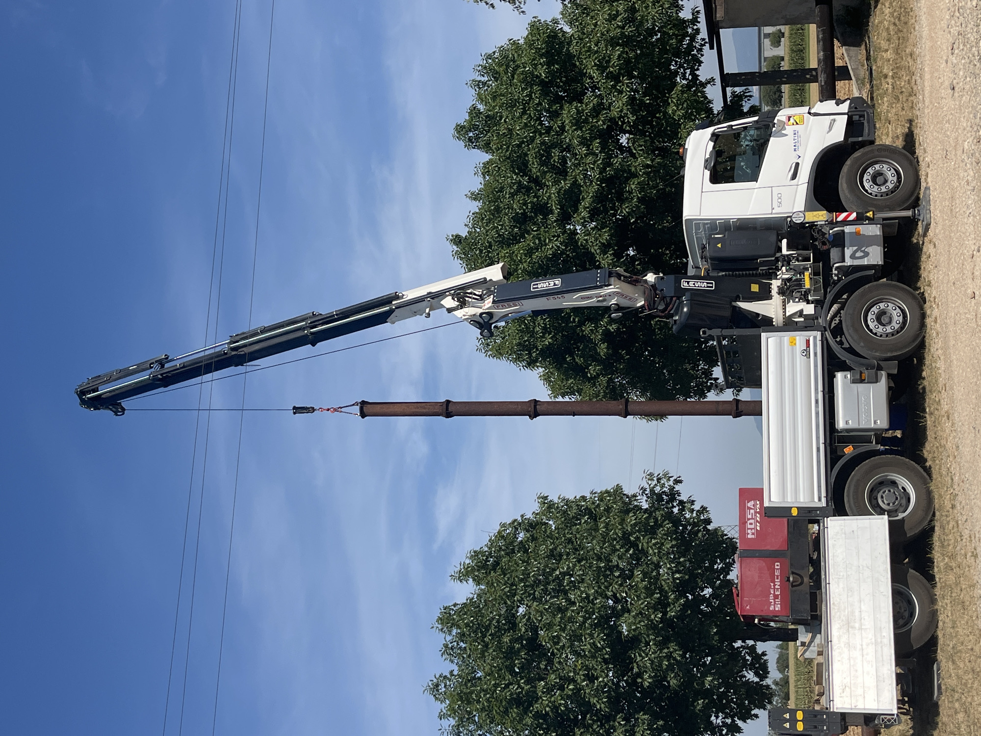 The image depicts a construction or demolition scene on a sunny day in Castiglione delle Stiviere, Italy. A white Volvo truck with a crane attachment is positioned on a gravel surface, with its outriggers deployed for stability. The crane is actively lifting a tall, rusty cylindrical object, presumably a pipe or a section of a chimney, into the air. The truck's flatbed carries a red, "SUPER SILENCED" generator. Large green trees flank the scene, and a clear blue sky with a few wispy clouds is visible above. Power lines are also present in the background. The overall environment suggests an outdoor industrial or infrastructure project. No people are directly visible in the image.