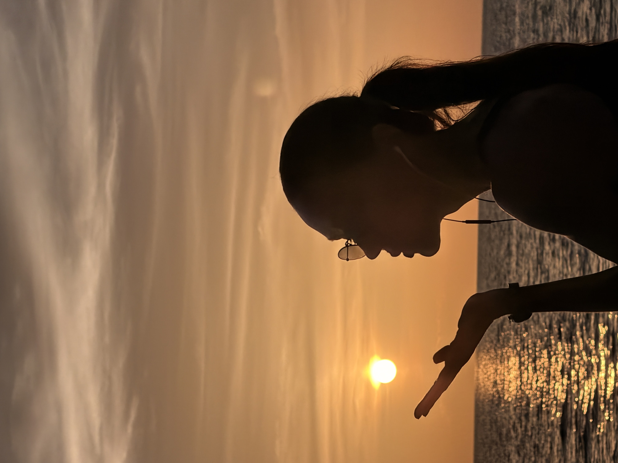 A silhouette of a woman wearing glasses and earphones stands by the sea during sunset in Ko Samui District, Thailand. The sun, a bright orb, is low on the horizon, casting a golden glow across the sky and the water. The woman's right hand is outstretched, as if catching the last rays of sunlight. Her hair is pulled back in a ponytail, and her profile is clearly visible against the vibrant orange and yellow hues of the sky. The ocean stretches out before her, its surface shimmering with reflected light. The overall mood is one of tranquility and natural beauty.