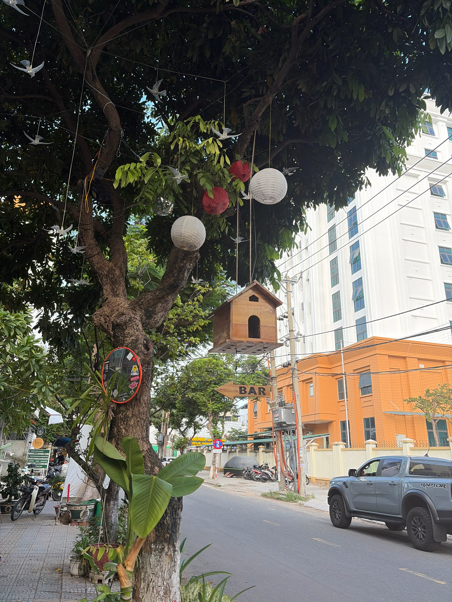 The image depicts a street scene in Đà Nẵng, Vietnam, with a focus on a tree adorned with decorations and a birdhouse. The tree, situated on the left side of the frame, is large and leafy, with several branches extending outwards. Hanging from the branches are numerous white and red paper lanterns, along with several decorative white bird figures. A small wooden birdhouse is also suspended from a branch.
On the tree trunk, a circular orange-framed mirror with stickers is attached. Various potted plants are visible at the base of the tree and along the sidewalk. To the right of the tree, a grey pickup truck, identified by "RAPTOR" badging, is parked on the street. Behind the truck, an orange building with several windows is prominent, and further in the background, a tall white building with a grid-like facade stands out. A wooden sign pointing to "BAR" is also visible on a pole near the orange building. The street itself is paved, and a sidewalk is present on the left. The overall impression is of a street in a city, likely in a commercial or residential area, with an artistic and decorative element introduced by the tree's embellishments.