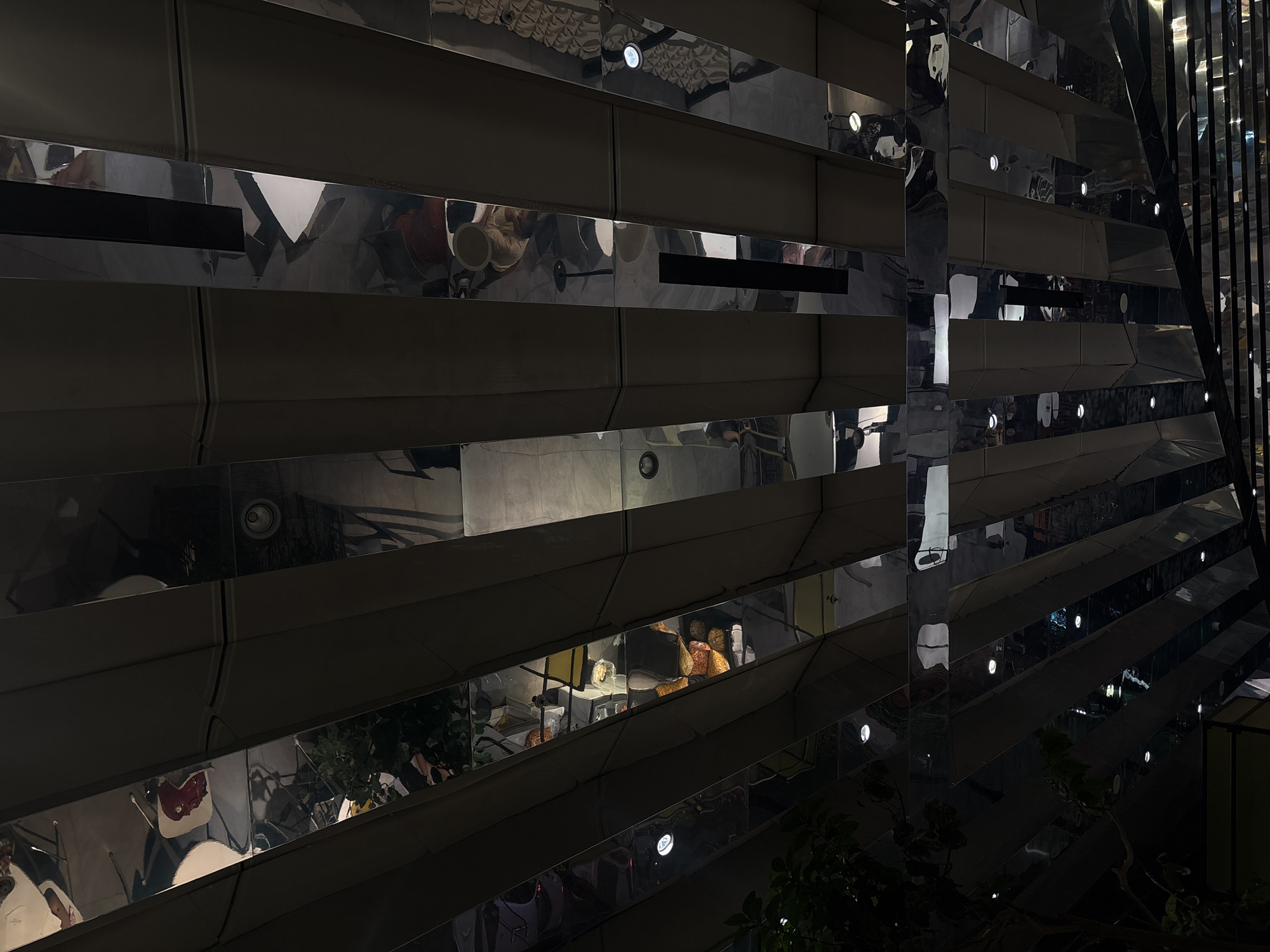 The image depicts the interior of a modern architectural space, likely a multi-story atrium or lobby in Mexico City, Mexico. The dominant feature is a series of horizontal, highly reflective, possibly mirrored or polished metal, bands that form a staggered or stepped pattern across the frame. These reflective surfaces create multiple, distorted reflections of the surrounding environment.
In the reflections, various elements of the building's interior are visible. The upper sections show reflections of a textured ceiling, featuring a patterned surface and numerous bright, circular recessed lights. Below this, reflections reveal parts of what appears to be a seating area, possibly a restaurant or lounge. These reflections include indistinct forms of people, white chairs, tables, and what could be a bar or display counter with objects that have orange-brown hues, possibly food items or decor. Some darker, rectangular openings, likely air vents, are also reflected in these bands.
Towards the bottom and to the left, reflections of green foliage indicate the presence of plants at a lower level. On the far right, a vertical reflective element, possibly a column or corner, continues the mirror-like effect, reflecting additional lights and the general interior.
The scene is dimly lit overall, with artificial light sources being the primary illumination. The bright reflections of the numerous circular lights suggest an evening or nighttime setting indoors. No specific activity is clearly discernible, but the reflections of people imply that the space is occupied and in use. The architectural style is sleek and contemporary, emphasizing geometric forms and reflective materials. There is no visible text in the image.