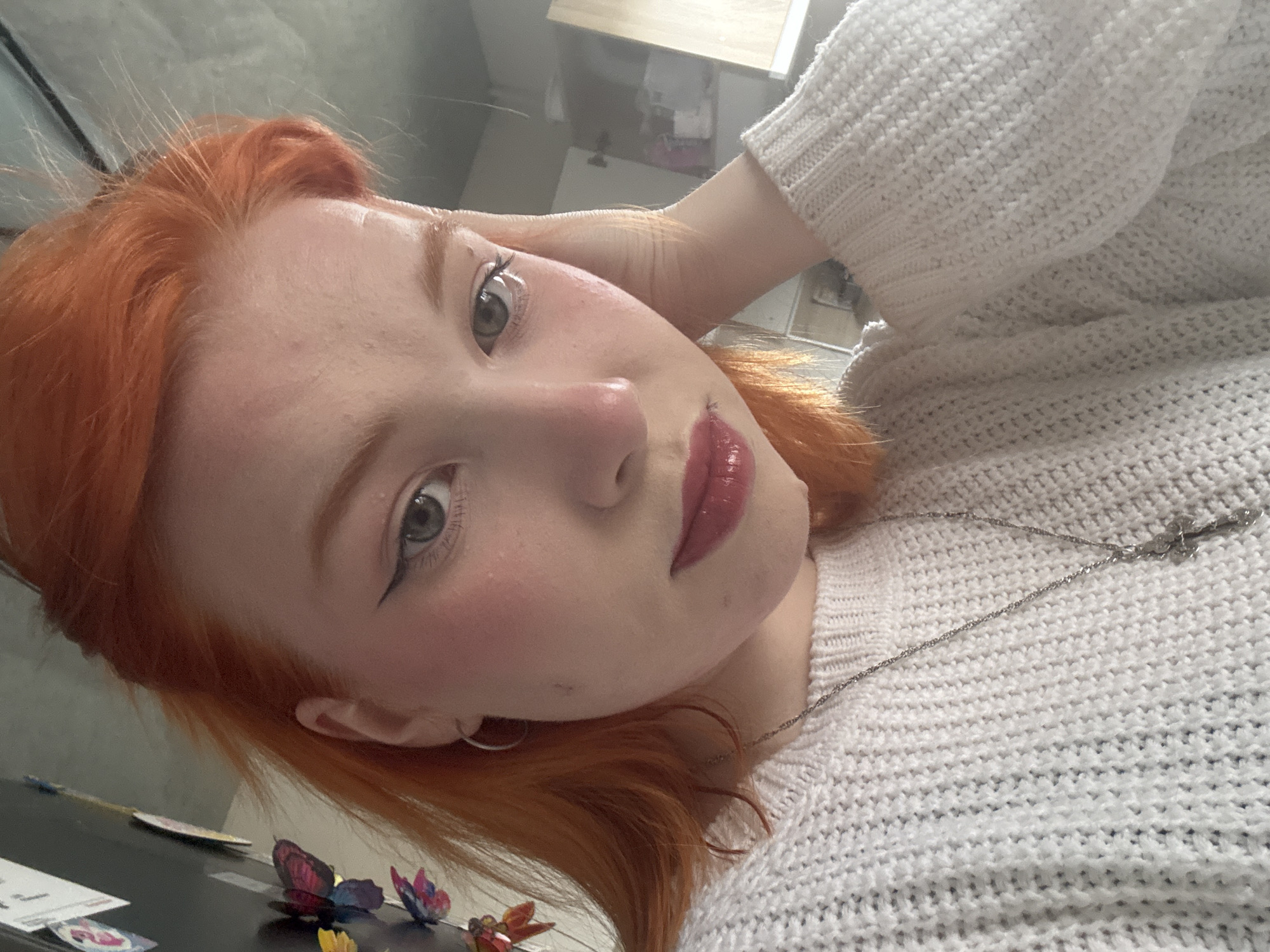 A close-up, tilted portrait captures a young person, likely female, with striking vibrant red-orange hair and fair skin. She looks directly at the camera with large, bright green-hazel eyes, featuring black winged eyeliner, a light blush on her cheeks, and glossy red lipstick. Her left hand is subtly raised, resting near the side of her head, gently pushing back her hair. She is wearing a cream or off-white knitted sweater, characterized by its textured weave, and a thin silver chain necklace. A small silver hoop earring is visible in her left ear. The background is softly blurred, revealing an indoor setting with reflections that suggest a room or office environment, possibly including a desk or table. In the lower left corner, a few small, decorative butterfly-shaped objects are visible on a dark surface. The lighting appears natural and bright, indicative of daytime. The person's expression is neutral and direct. No discernible text is visible.