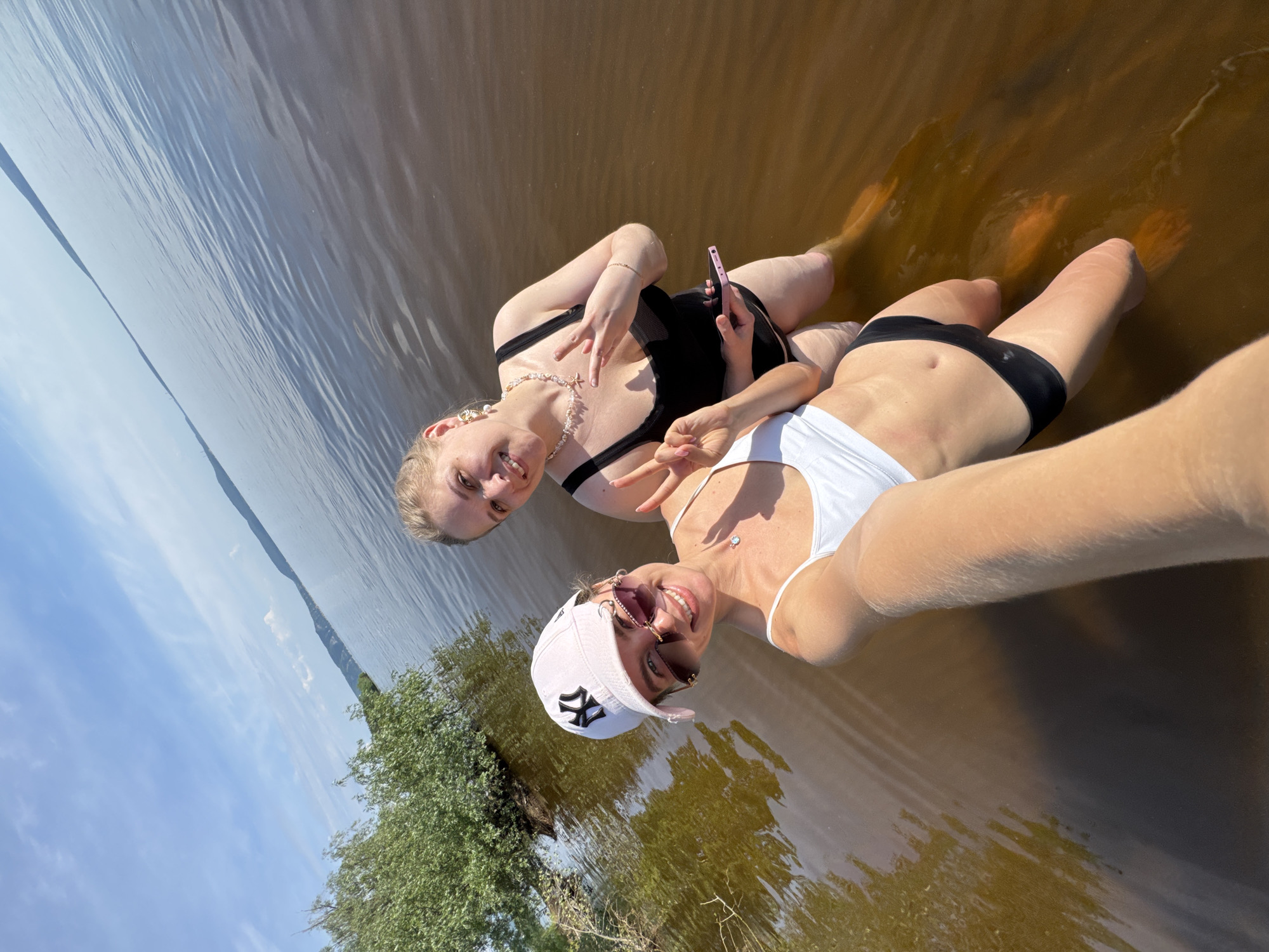 This is a selfie taken by two young women wading in shallow, murky water. The woman in the foreground is wearing a white bikini top and a pink baseball cap with a black "NY" logo. She is smiling and wearing sunglasses. The woman behind her is wearing a black swimsuit and has blonde hair. She is also smiling and holding up a cell phone. Both women are making peace signs with their fingers. The water appears to be a lake or river, with trees visible on the shore in the background. The sky is blue with some white clouds, suggesting it is a sunny day. The water is reflecting the sunlight, creating a shimmering effect. The overall mood of the image is casual and joyful, suggesting a fun summer outing.