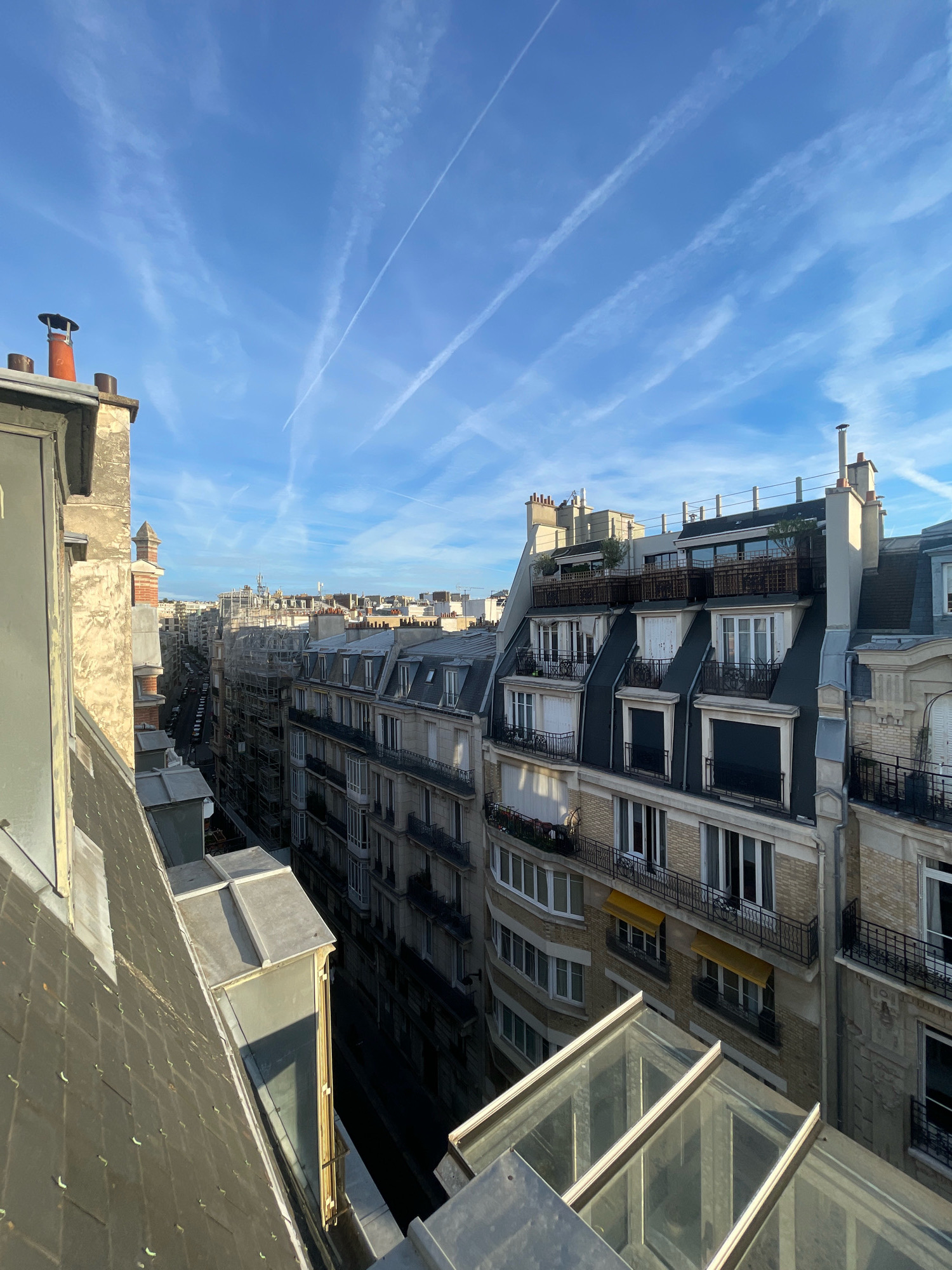 The image captures a high-angle view looking down a street in Paris, France, with several buildings lining both sides. The sky is a clear blue with scattered white contrails from airplanes. In the foreground, a rooftop with a sloped, grey tiled surface is visible, along with some skylight structures and the side of a building.
The buildings on either side of the street are tall, multi-story structures typical of Parisian architecture, featuring mansard roofs with dormer windows. Many of these windows have black wrought-iron balconies. Some buildings have a yellowish brick facade, while others appear to be made of stone. Several buildings show signs of ongoing renovation, with scaffolding visible on one. The street below is lined with parked cars, and the sidewalks are narrow.
The overall impression is one of an urban cityscape on a clear day, showcasing the characteristic architectural style of Paris. There are no people or animals visible in the image.