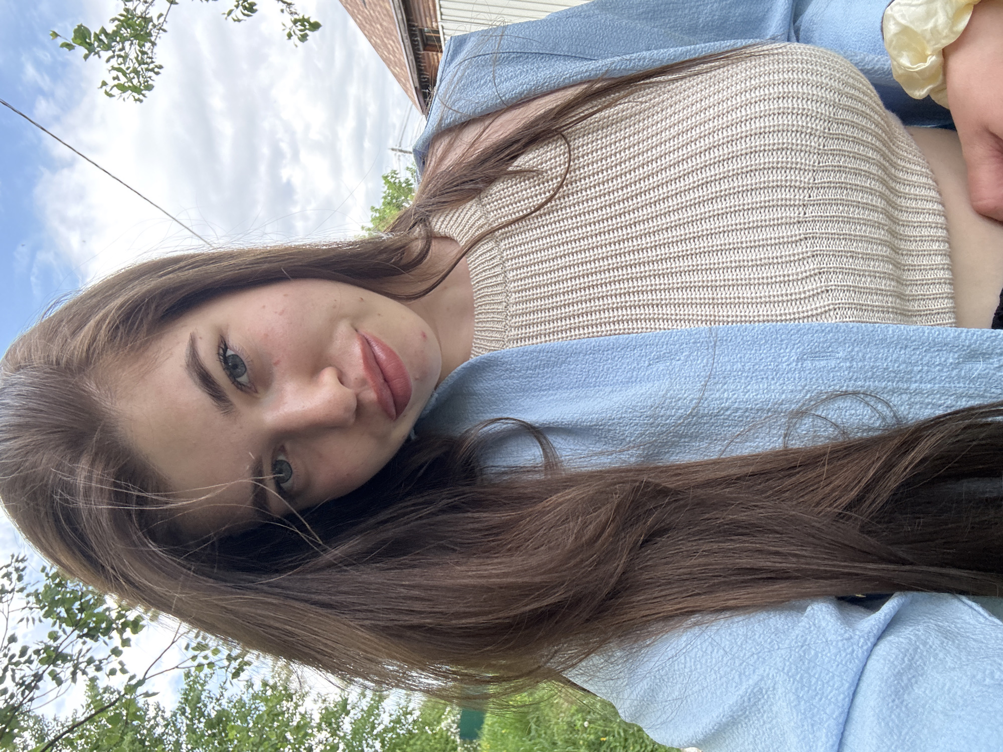 A woman with long brown hair, wearing a light blue jacket over a cream-colored knitted top, is posing outdoors. The background includes a sky with clouds, trees, and a building with a brown roof. The location is ambiguous, and there is no information to suggest that the location is Мещёры, Russia.