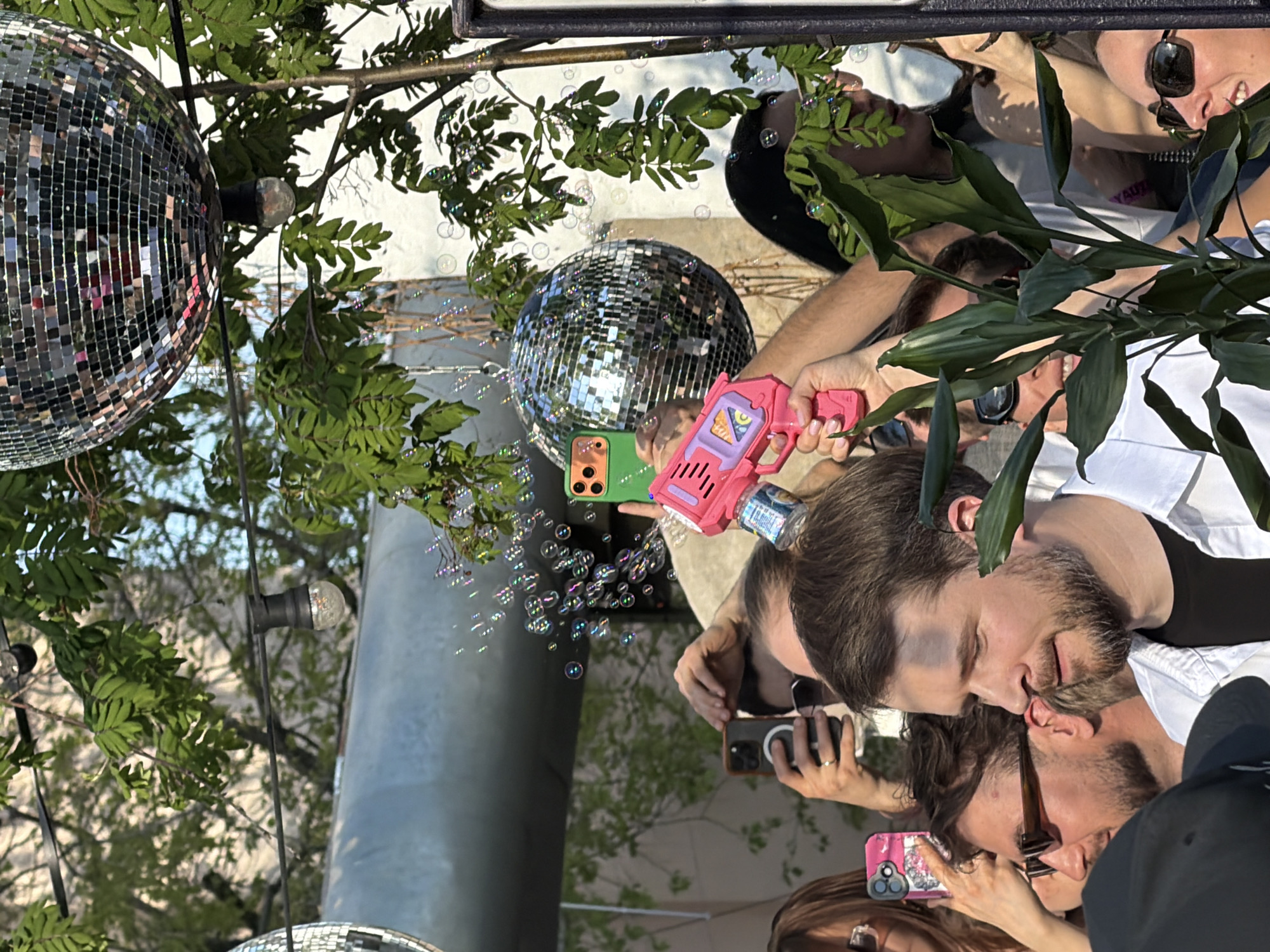 The image shows a group of people gathered outdoors, possibly in a public space, with disco balls hanging overhead amidst greenery, possibly in Moscow, Russia. People are taking photos and videos, with one person holding a pink bubble gun, producing bubbles visible in the air. The background includes a cylindrical structure, suggesting an urban setting. The people are dressed in casual clothing, and the overall atmosphere appears festive and social.