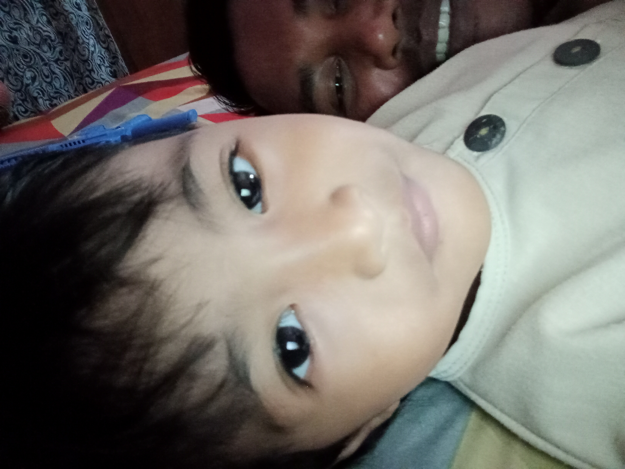 A low-angle, slightly rotated, close-up photograph shows two people lying down, possibly in a bed. The person in the foreground is a child, whose face is turned towards the camera. The child has dark hair, dark eyes, and fair skin. The child is wearing a light-colored garment with two dark circular buttons. The person in the background is an adult, partially visible behind the child, with dark hair and a smiling expression. The background shows bedding with a striped pattern and a dark, patterned fabric. The scene appears to be indoors. The location is Bloro, Indonesia.