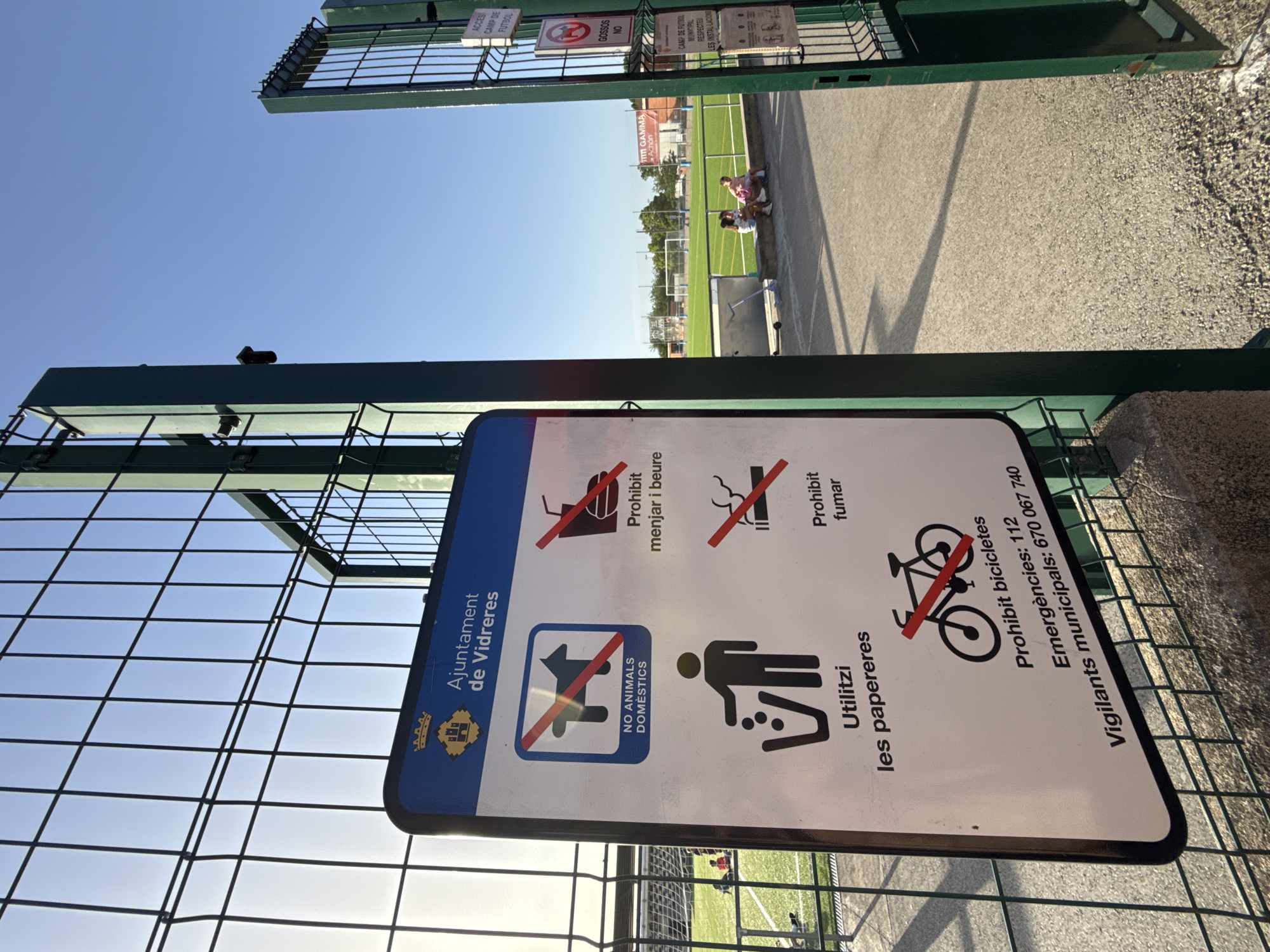 A close-up, eye-level shot captures a metal fence and a sign at the entrance to the Camp de Futbol Municipal in Vidreres, Spain. The sign, predominantly white with a blue border, displays several regulations in both Catalan and English, including prohibitions against domestic animals, eating and drinking, smoking, and cycling. It also instructs visitors to use the trash cans and provides emergency contact numbers. The sky is clear and blue. In the background, a well-maintained artificial turf soccer field is visible, with goalposts at either end. A few people are seated on a low wall near the field, and a scooter lies on the ground. Another sign on a green metal post to the right of the frame, also in Catalan, indicates "ACCÉS CAMP DE FUTBOL" and lists regulations, including "GOSSOS NO," meaning "dogs no."