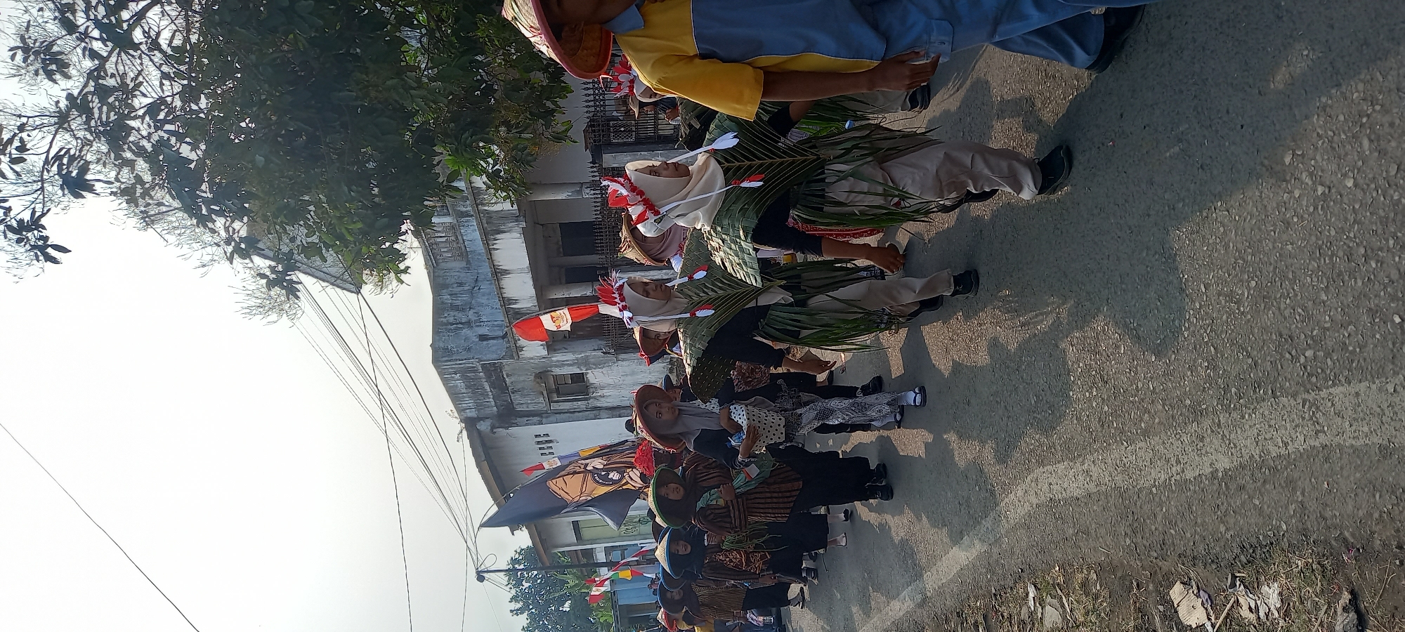 A group of people, primarily young women, are parading down a paved street in Wanareja, Indonesia, under a bright, hazy sky. They are dressed in traditional attire, with many wearing conical straw hats and patterned brown and white long-sleeved shirts and pants. Some are adorned with elaborate costumes made of palm fronds, resembling wings or capes. They are decorated with red and white feathers on their heads, reminiscent of Indonesian flags. One person in the foreground on the right wears a yellow and blue collared shirt and blue pants. The parade appears to be part of a cultural event or festival. In the background, a weathered building with a barred fence and a flagpole displaying flags is visible. Overhead, power lines crisscross the sky. The street is lined with dry grass and debris on the left side.