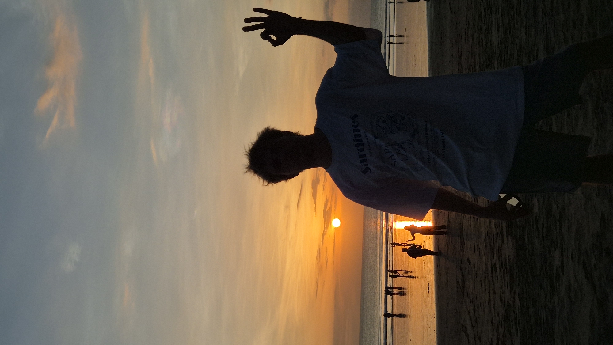 A vertical outdoor shot depicts a person silhouetted against a sunset on a beach, likely in Kuta, Indonesia. The figure is standing and making a hand gesture with fingers forming a small circle. The person wears a light-colored short-sleeved shirt with possible text or design on it, and dark shorts. The sunset casts an orange glow across the sky and sea, with the sun appearing as a bright orb on the horizon. Other people are visible as silhouettes along the shoreline. The beach is dark in the foreground. The sky is tinted with a mix of orange and gray shades. The photo's orientation is turned sideways.